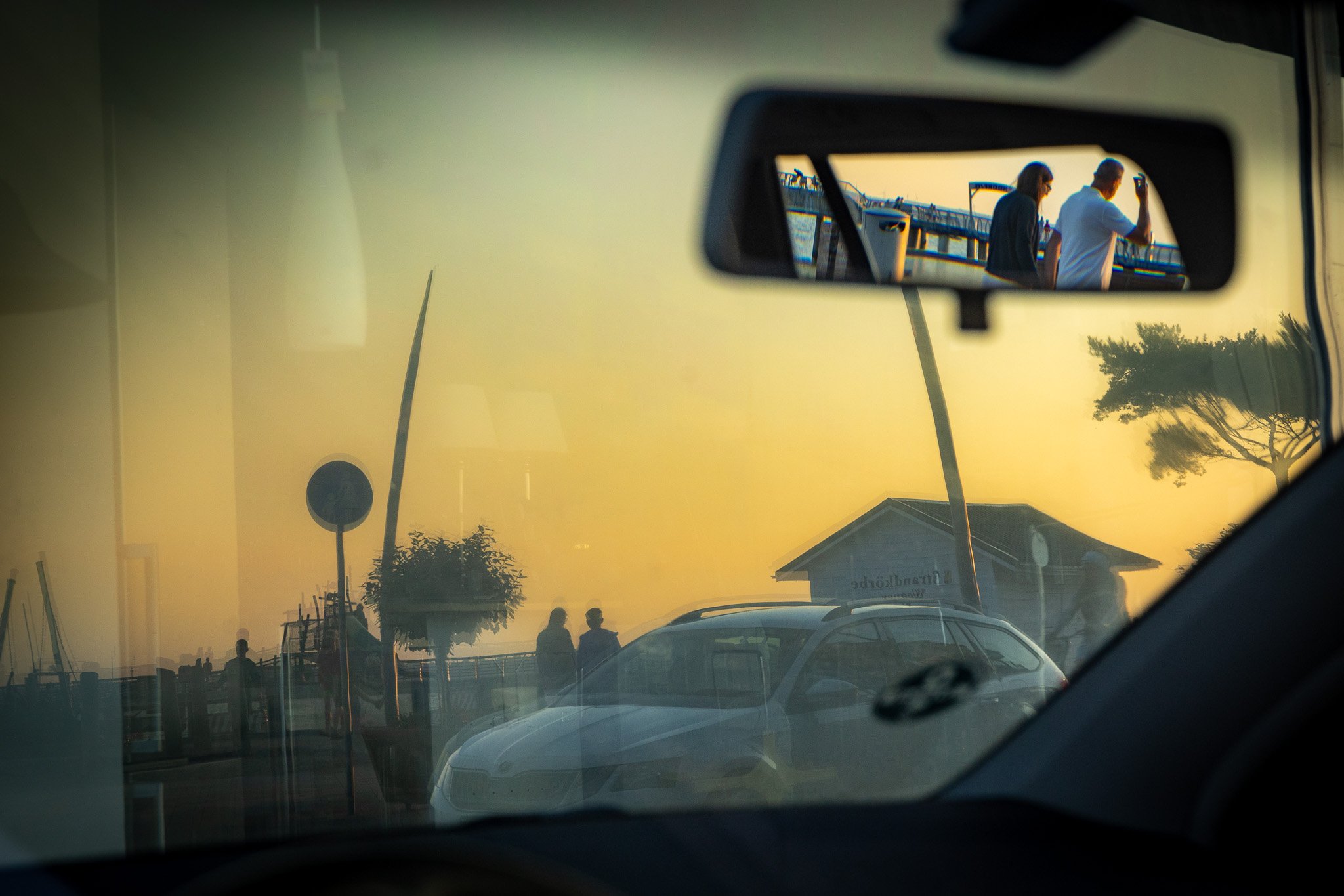 Blick durch die Windschutzscheibe eines Autos, mit zwei Personen auf einer Brücke bei Sonnenuntergang, umgeben von geparkten Autos, einem Baum und Gebäuden im Hintergrund.