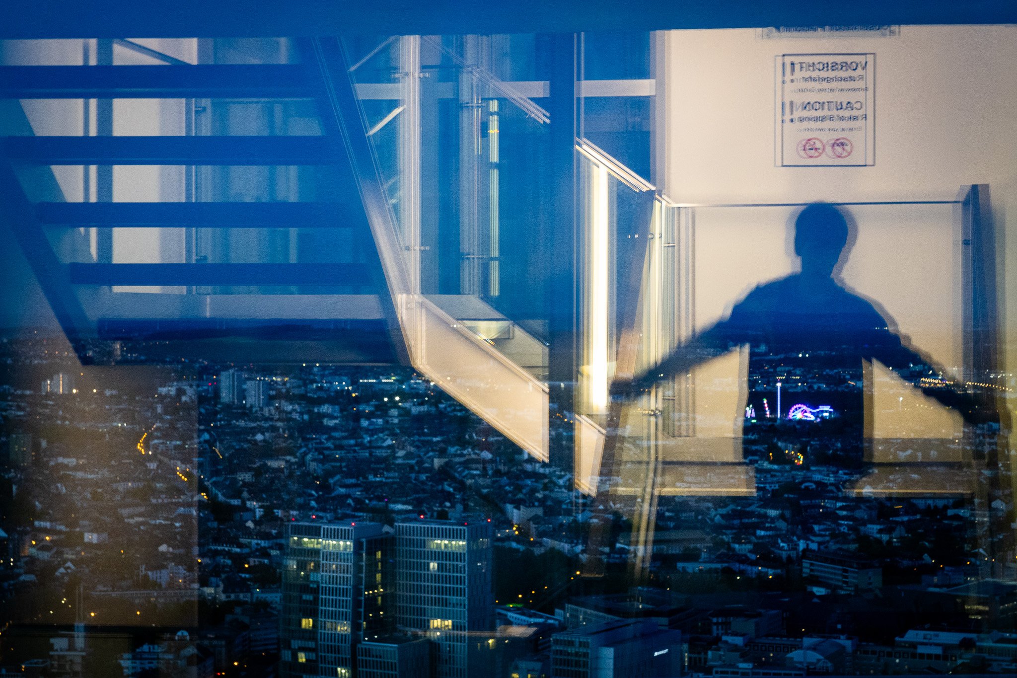 Ein Blick durch eine Fensterscheibe auf eine Stadt bei Nacht mit beleuchteten Gebäuden und einem Vergnügungspark im Hintergrund sowie dem Schatten einer Person, die im Inneren des Gebäudes steht.