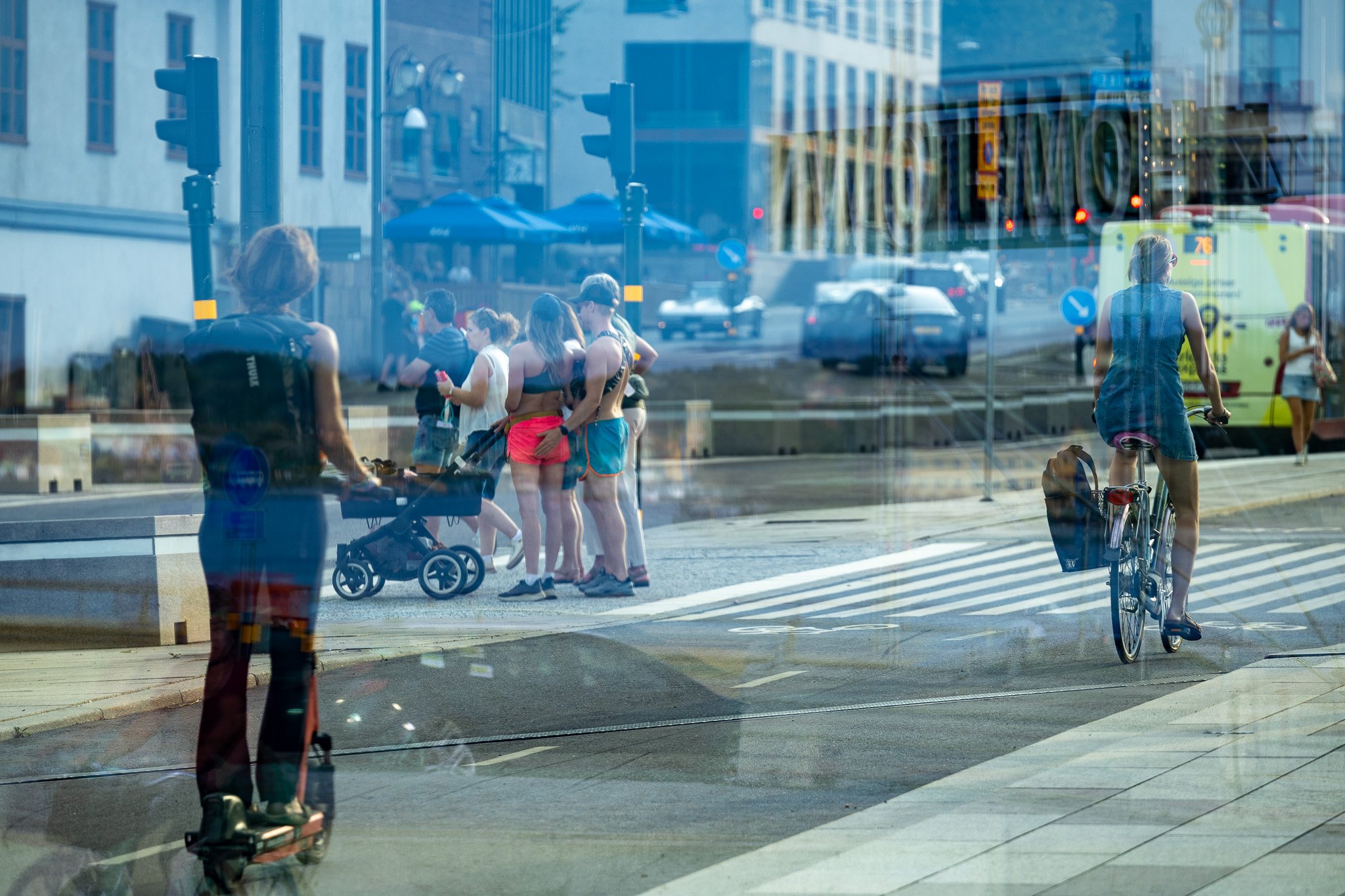 Am Straßenrand stehen Menschen, einige mit Kinderwagen, und Radfahrer warten darauf, die Straße überqueren zu können. Im Hintergrund sind Gebäude, ein Bus und ein Auto zu sehen. Das Bild wurde durch eine reflektierende Glasscheibe aufgenommen.