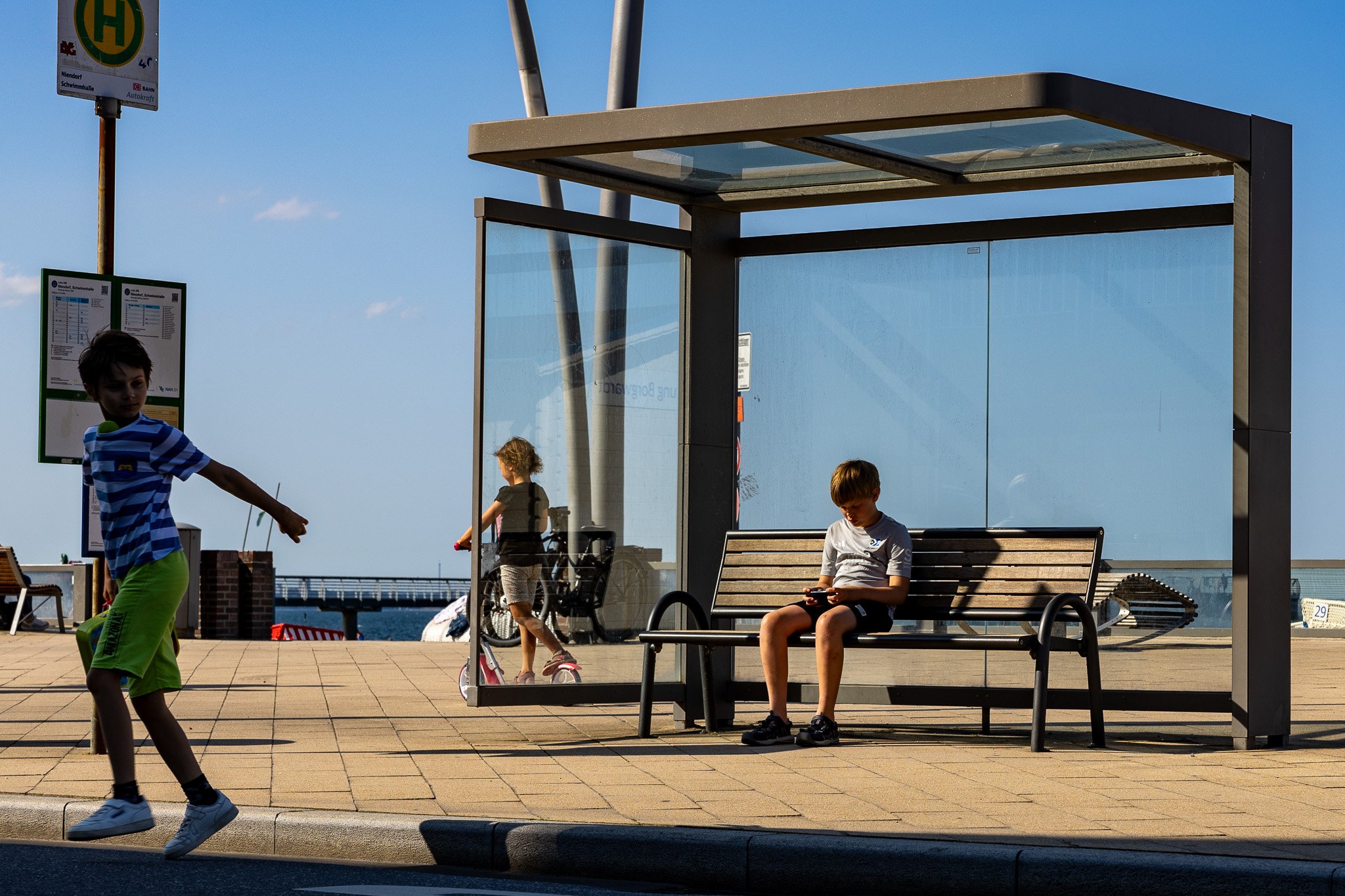 Menschen an einer Bushaltestelle, ein Junge läuft vorbei, ein Junge sitzt auf einer Bank und schaut auf sein Smartphone, ein Mädchen steht neben einem Fahrrad, im Hintergrund eine Brücke und Wasser, der Himmel ist blau mit wenigen Wolken.