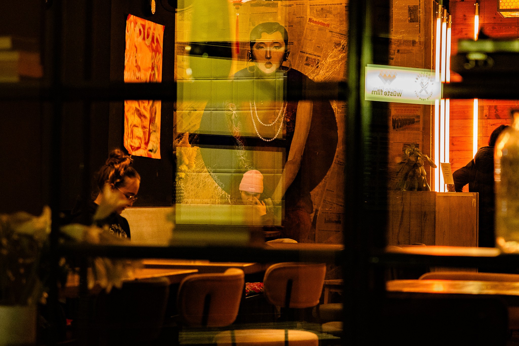 Blick durch ein Fenster in ein Lokal bei Nacht, mit einigen Gästen und einer Frau in schwarzer Kleidung, die im Hintergrund auf einer Leinwand zu sehen ist.