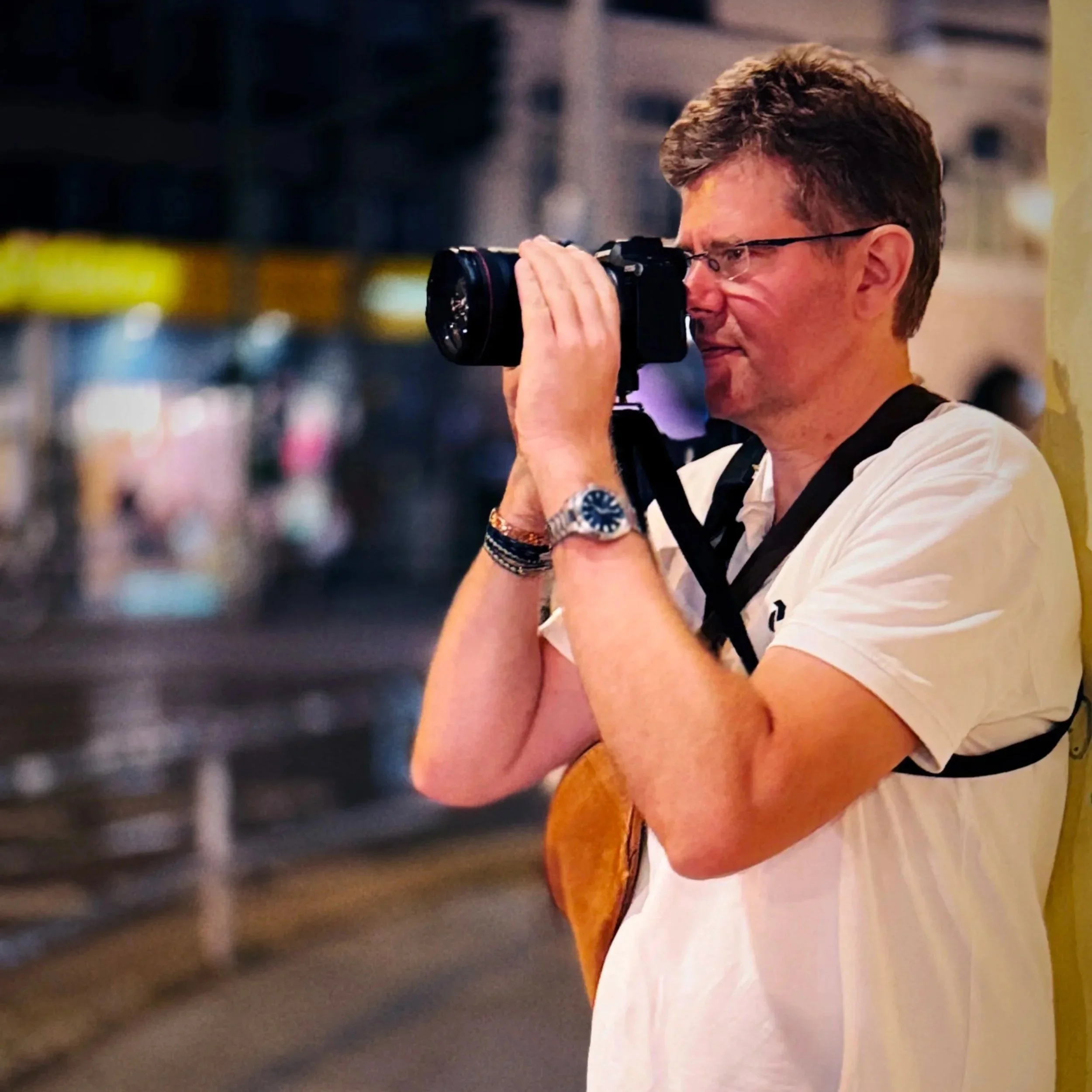 Ein Mann mit Brille und Armband fotografiert eine Szene mit einer Kamera; im Hintergrund sind unscharfe Lichter und Personen zu sehen.