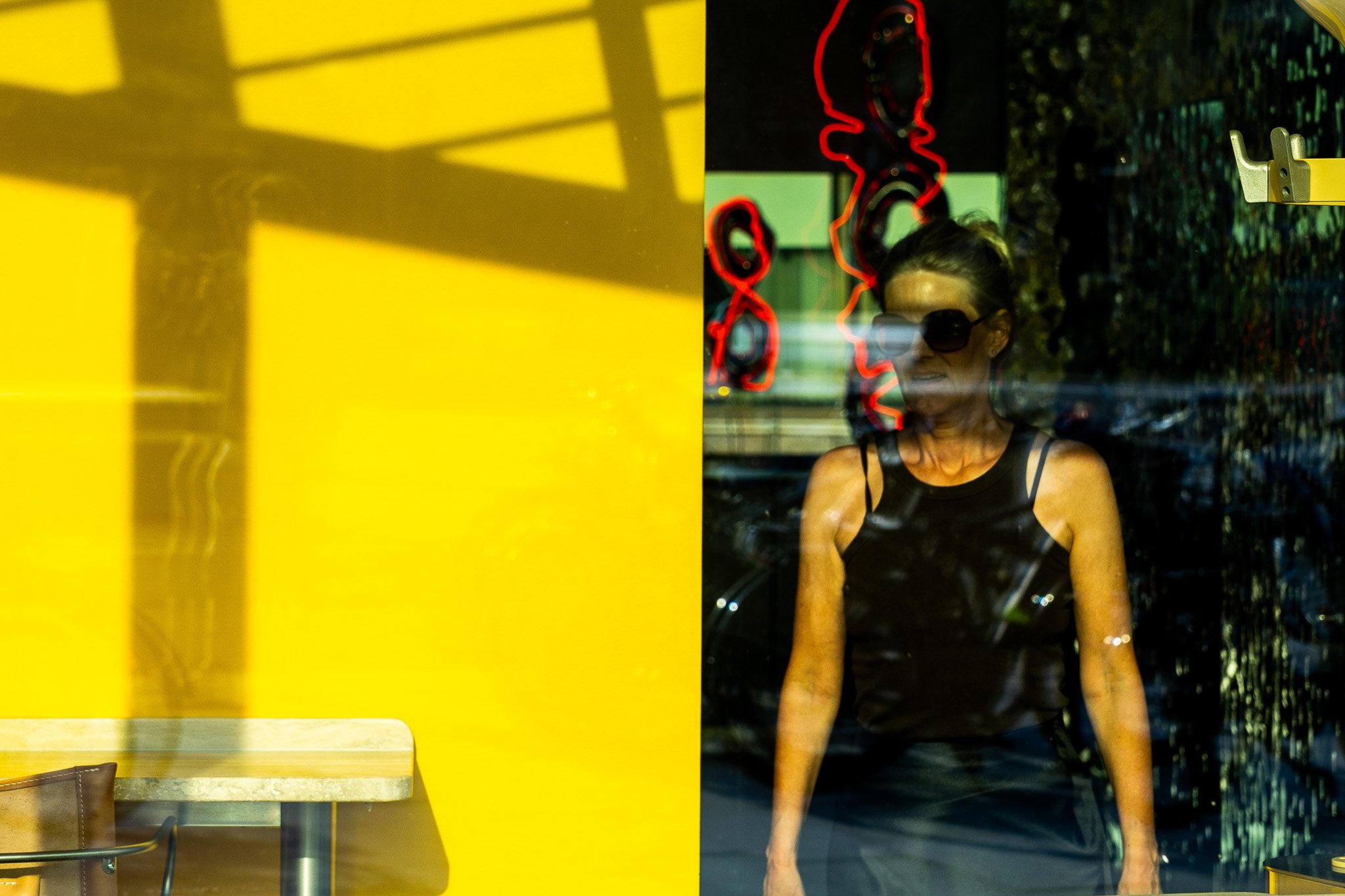 Ein Mann trägt ein schwarzes Tanktop, sitzt hinter einer Glasscheibe und betrachtet Neonringe an der Wand. Das Bild wird auf beiden Seiten von Fenstern eingerahmt, wobei auf der linken Seite eine gelbe Wand und eine Tischplatte zu sehen sind.