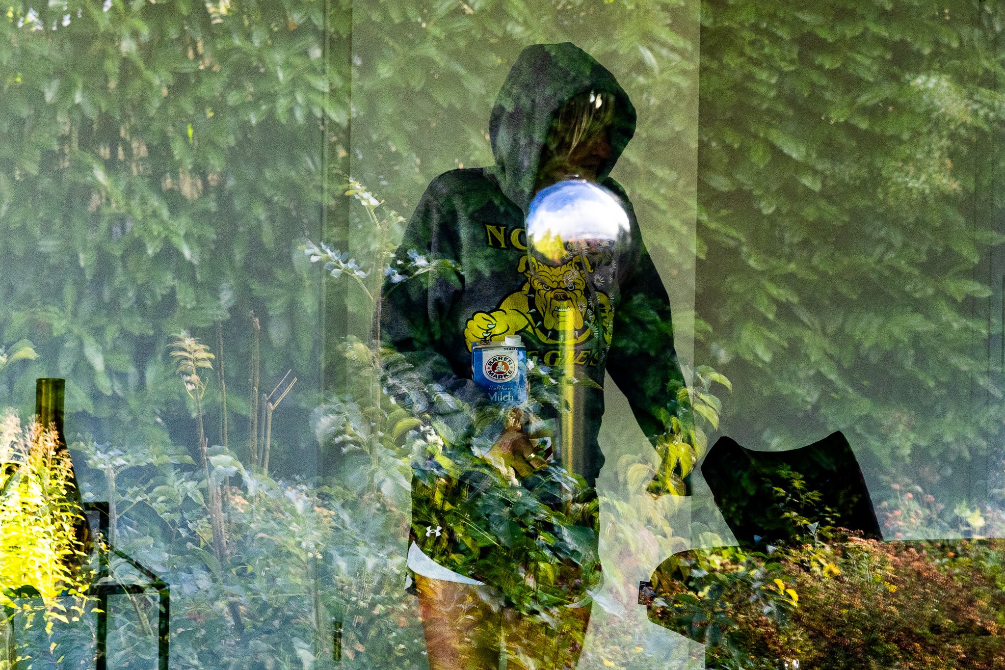 Eine Person mit Kapuzenpulli sitzt hinter einer Glasscheibe im Garten, umgeben von Pflanzen und Bäumen, und hält eine Flasche in der Hand.