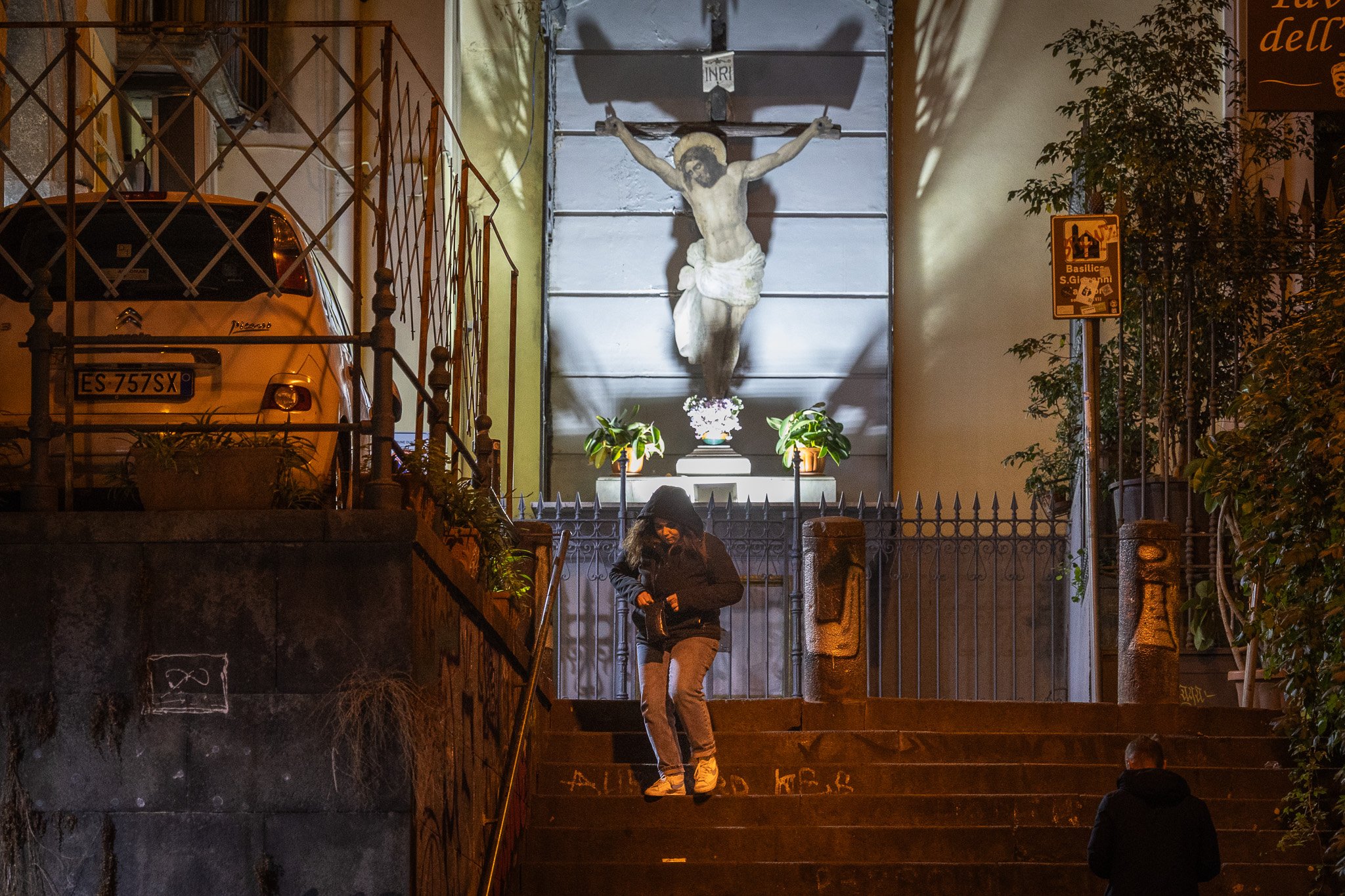 Eine Statue von Jesus am Kreuz in einer nächtlichen Szene, geprägt von warmem Licht, umgeben von Pflanzen, mit einer Person, die die Treppe hinuntergeht.