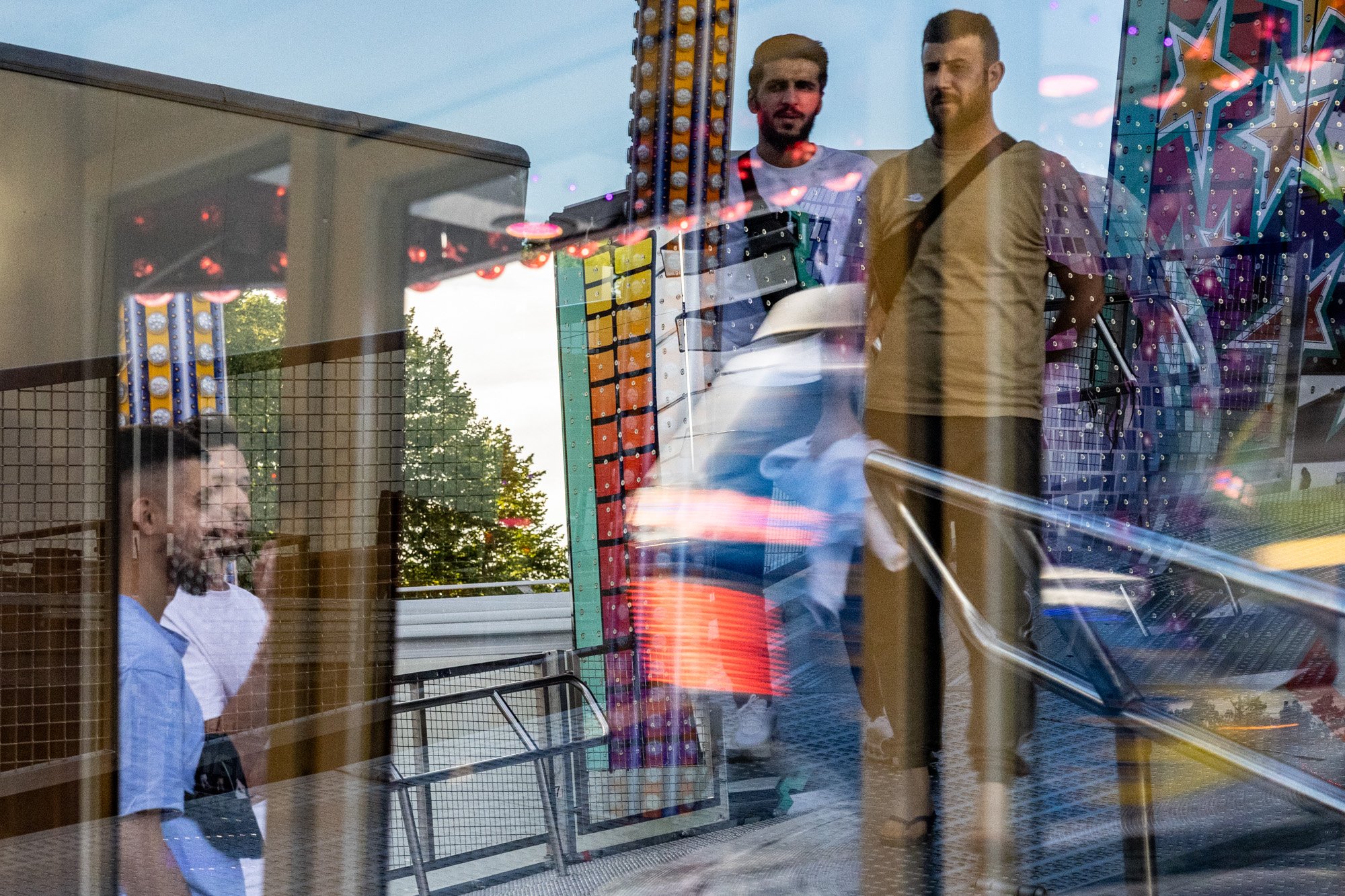 Spiegelbild von Menschen, die an einem Fahrgeschäft auf einem Jahrmarkt stehen, in einer Glasscheibe. Im Hintergrund sind bunte Lichtdekorationen zu sehen.