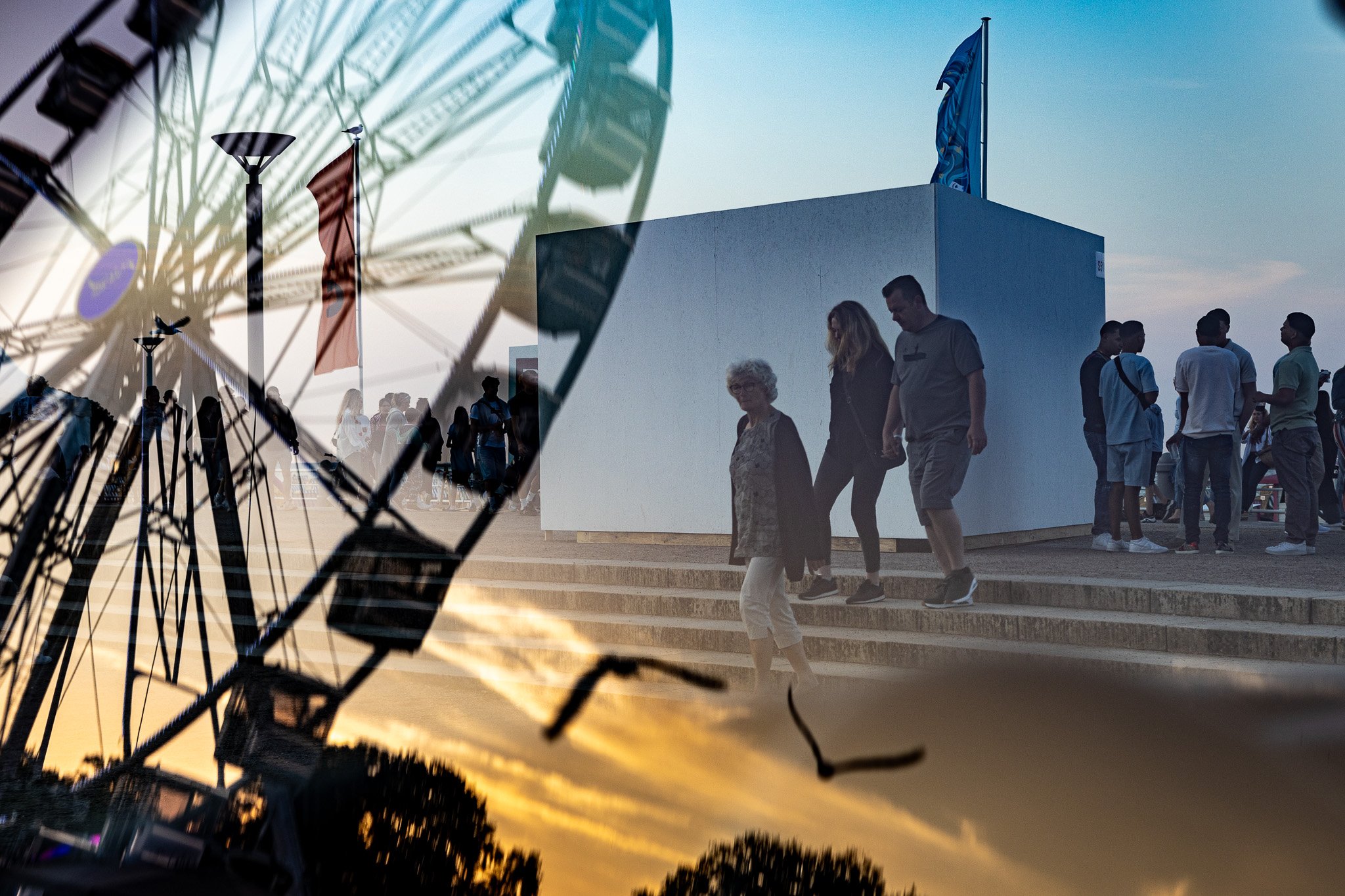 Doppelbelichtetes Foto eines Riesenrads bei Sonnenuntergang, auf dem Menschen eine Treppe hinaufgehen und ein weißes Gebäude mit einer blauen Flagge auf dem Dach zu sehen ist.