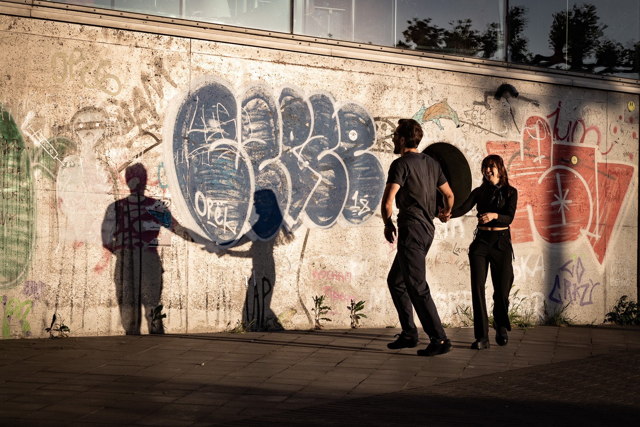 Zwei Menschen bei Sonnenuntergang, die vor einer mit Graffiti besprühten Wand stehen – eine Frau und ein Mann, die sich an den Händen halten und lachen.