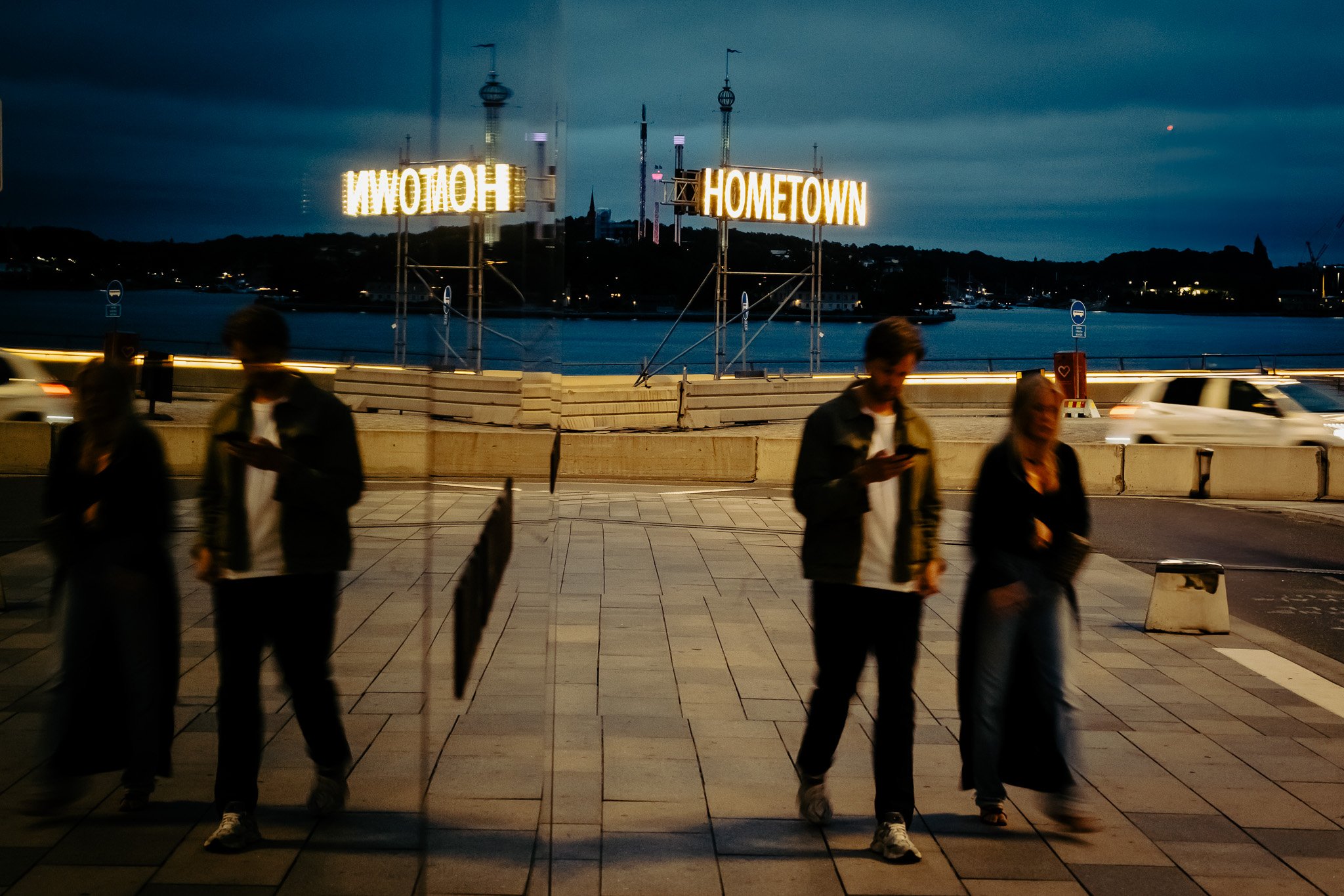 Blick auf eine Brücke am Abend mit einem beleuchteten Schild, auf dem „HOMETOWN“ steht, und Menschen, die vorbeigehen; im Hintergrund ein Gewässer mit Häusern und Wäldern.