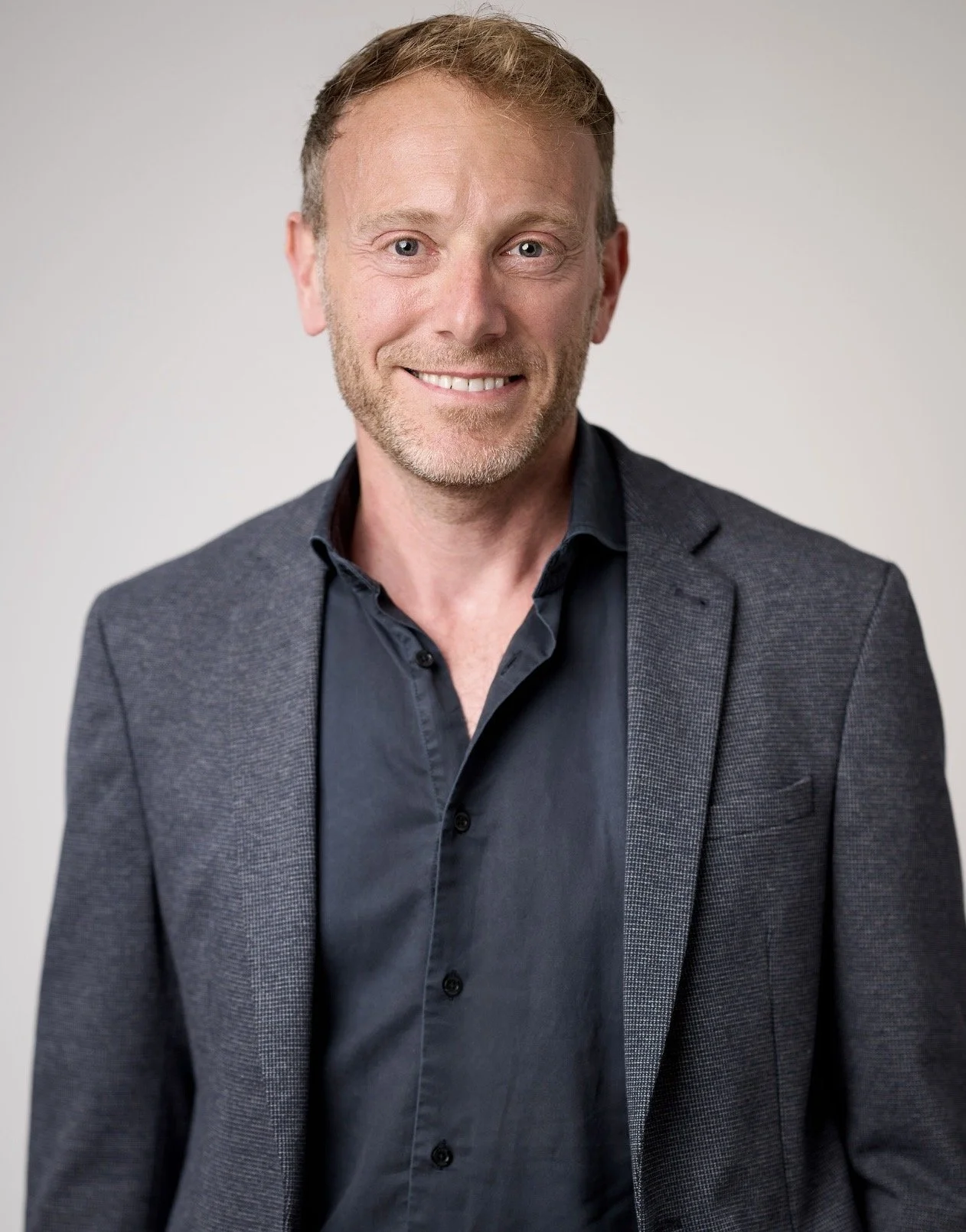 A portrait of a smiling man with light brown hair, light beard, wearing a dark blue shirt and a gray blazer, standing against a plain light background.