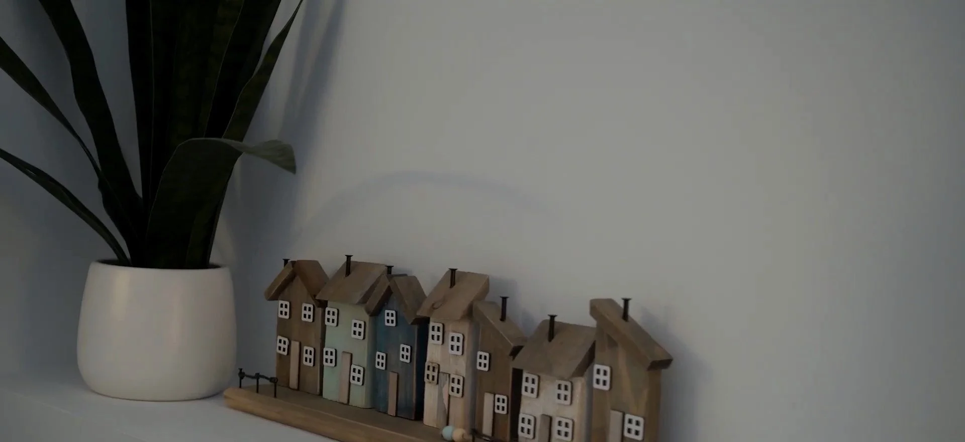 Indoor decor with a white planter holding a large leafy green plant and a row of small decorative wooden houses with miniature chimneys on a wooden base against a plain white wall.