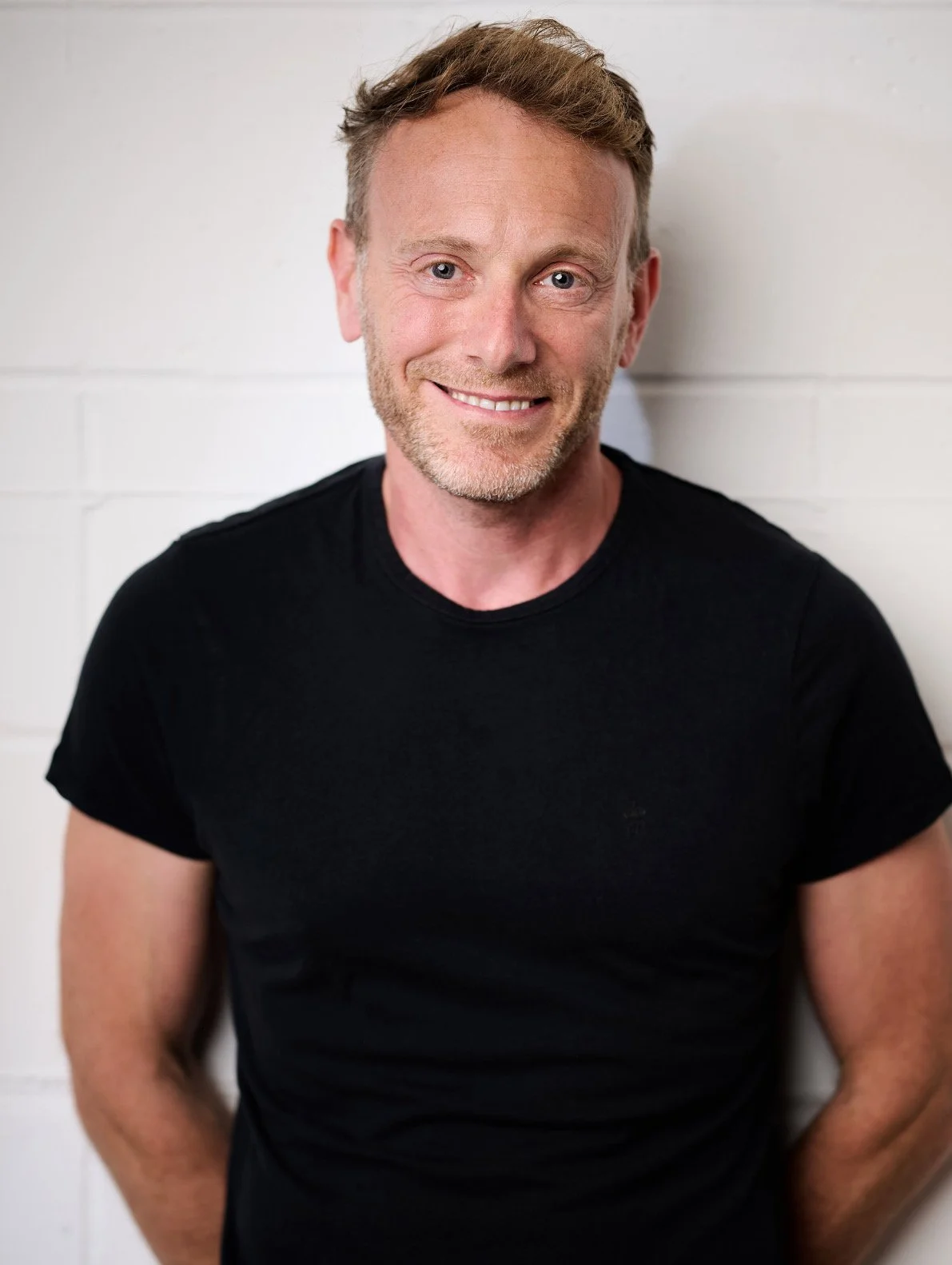 A smiling man with short light brown hair and a beard wearing a black t-shirt, standing against a white brick wall.