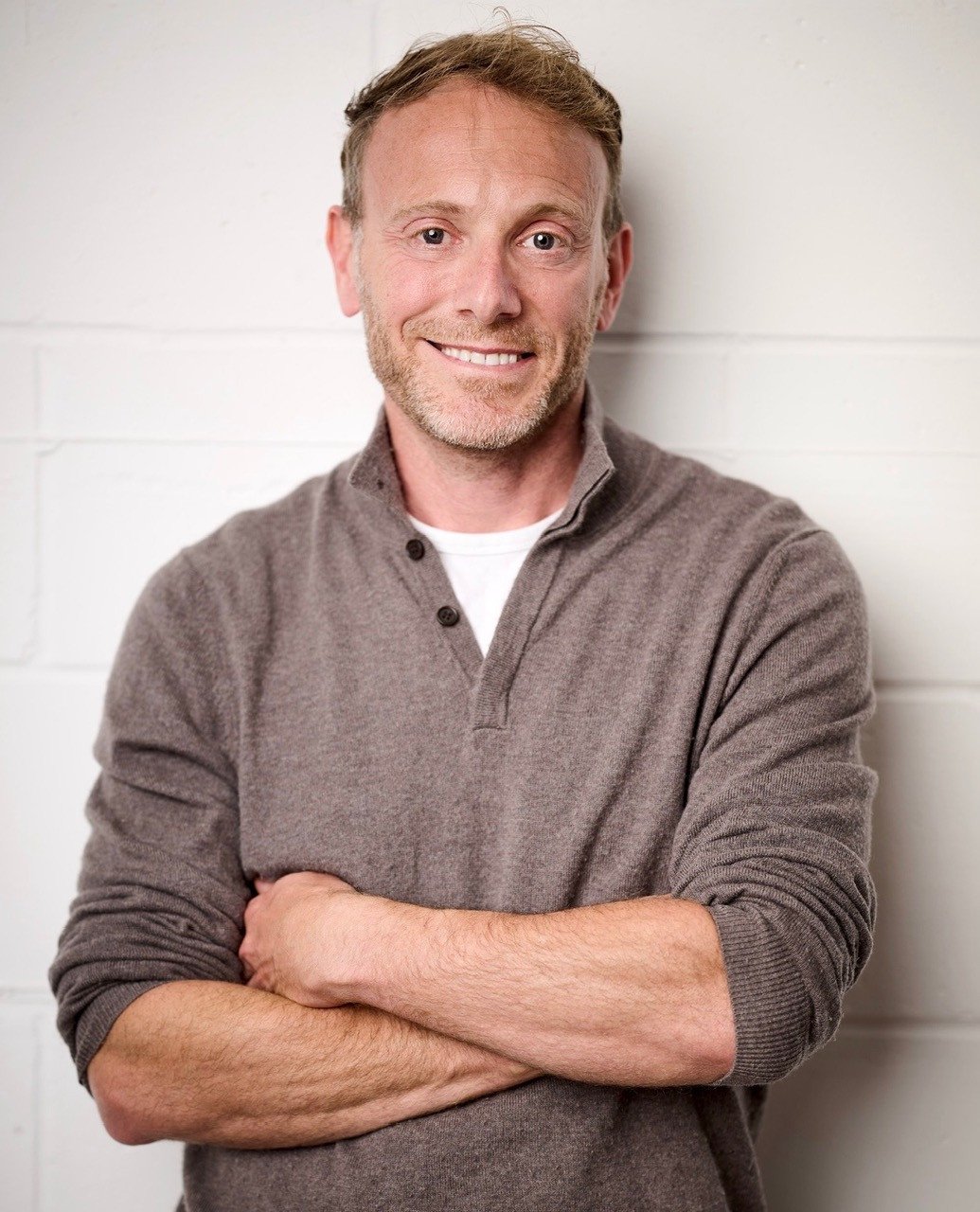 A smiling man with short, light brown hair and a beard, wearing a brown sweater and white shirt, stands with arms crossed against a white brick wall.