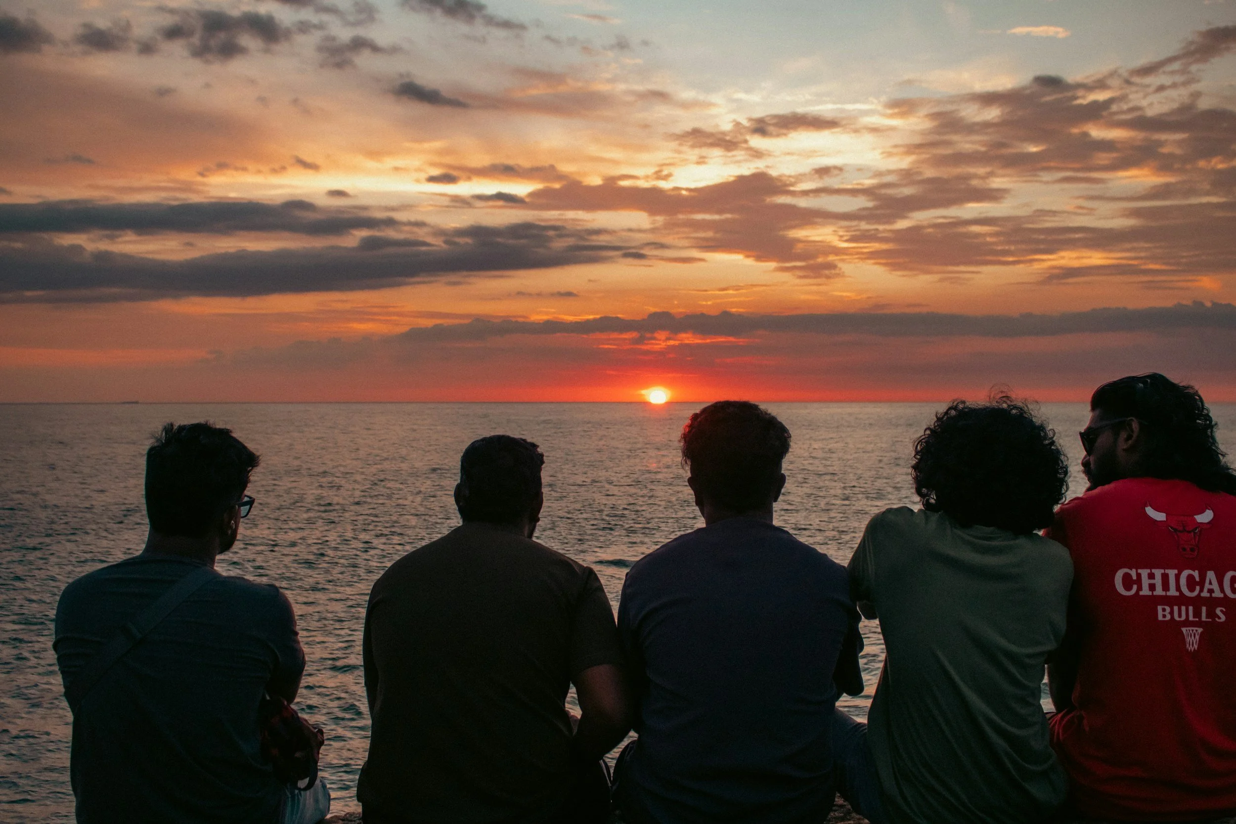Five friends sitting by the water watching a sunset with colorful clouds in the sky.
