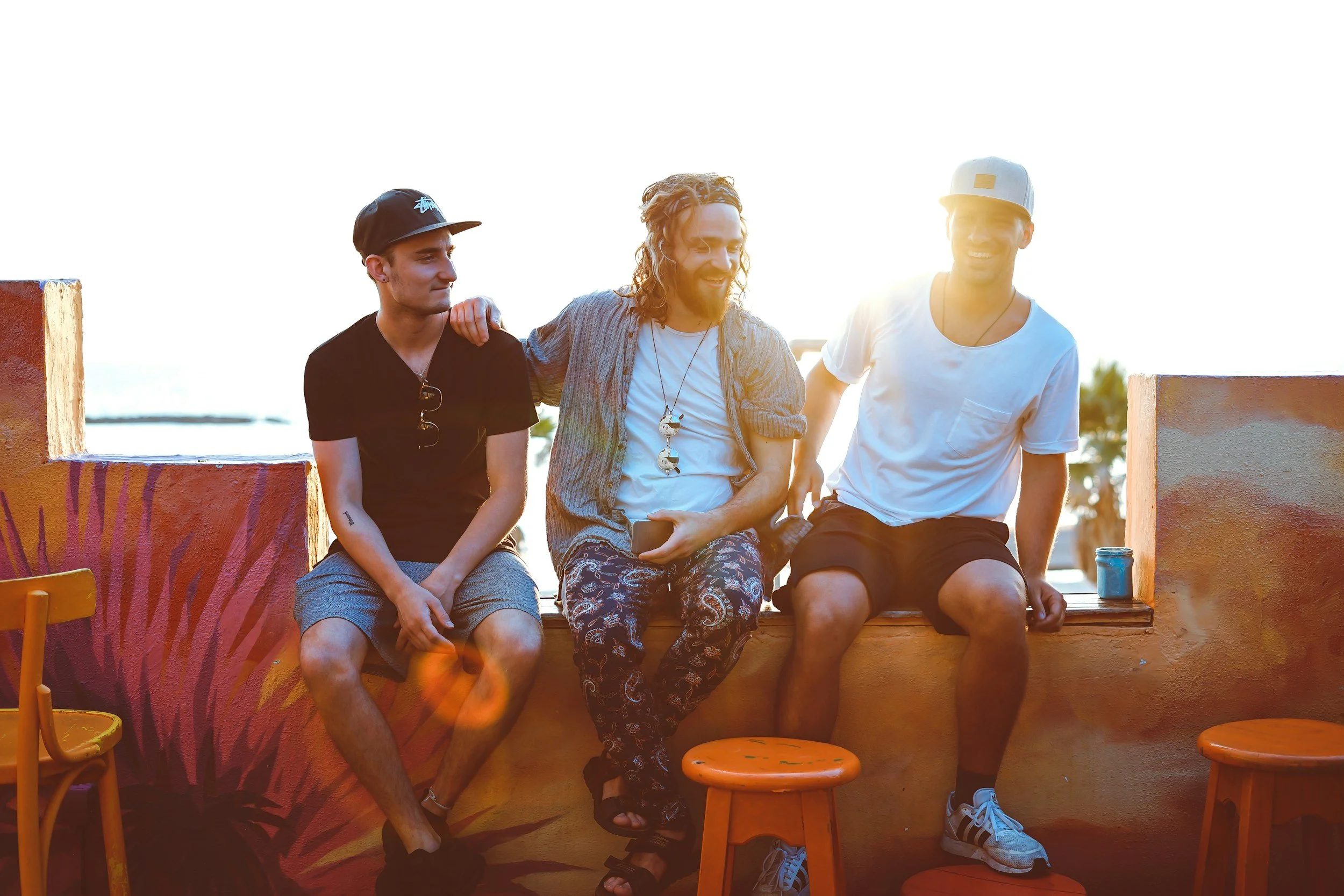 Three young men sitting on a wall outdoors, with one resting his arm on another's shoulder, all smiling and relaxed, with sunlight behind them.