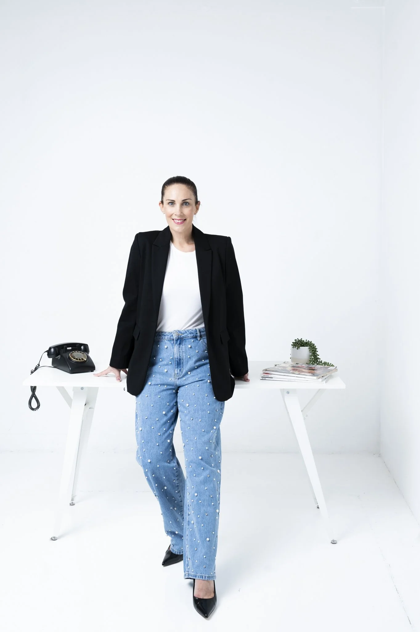 A woman with dark hair tied back, wearing a black blazer, white top, and blue jeans with pearl embellishments, stands in a minimalist white room. She is smiling, positioned in front of a white table with a vintage telephone, some magazines, and a sma