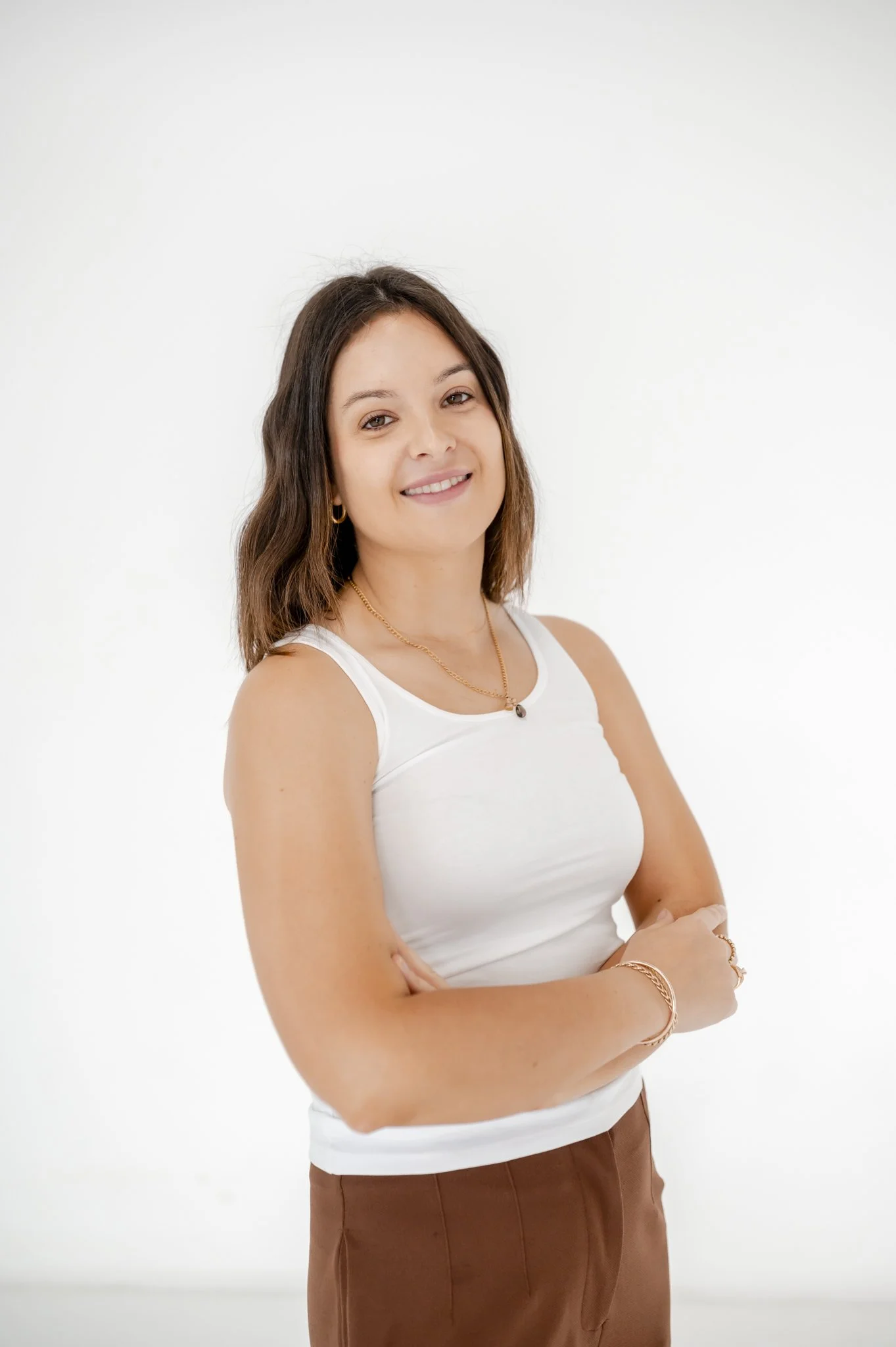 Portrait of a young woman with brown hair, wearing a white tank top, gold jewelry, and smiling against a plain white background.