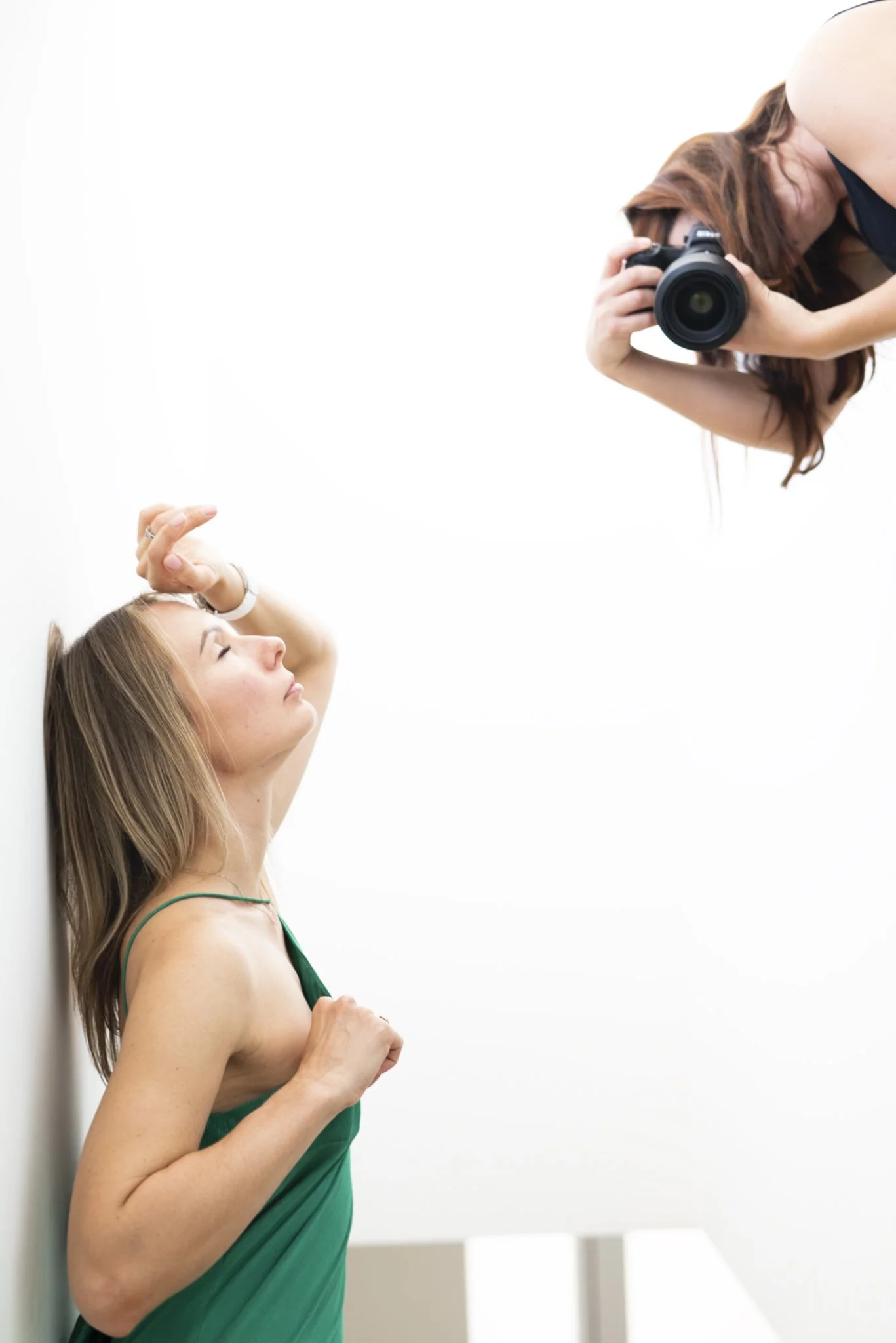 Woman in a green dress with long hair, leaning against a white wall, with her eyes closed and hand resting on her head, while another woman takes a photo of her with a camera.