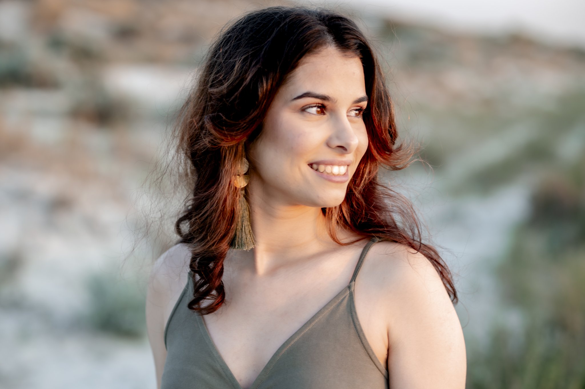 A young woman with long wavy brown hair with highlights, smiling and looking to her right, outdoors near the water with blurred background.
