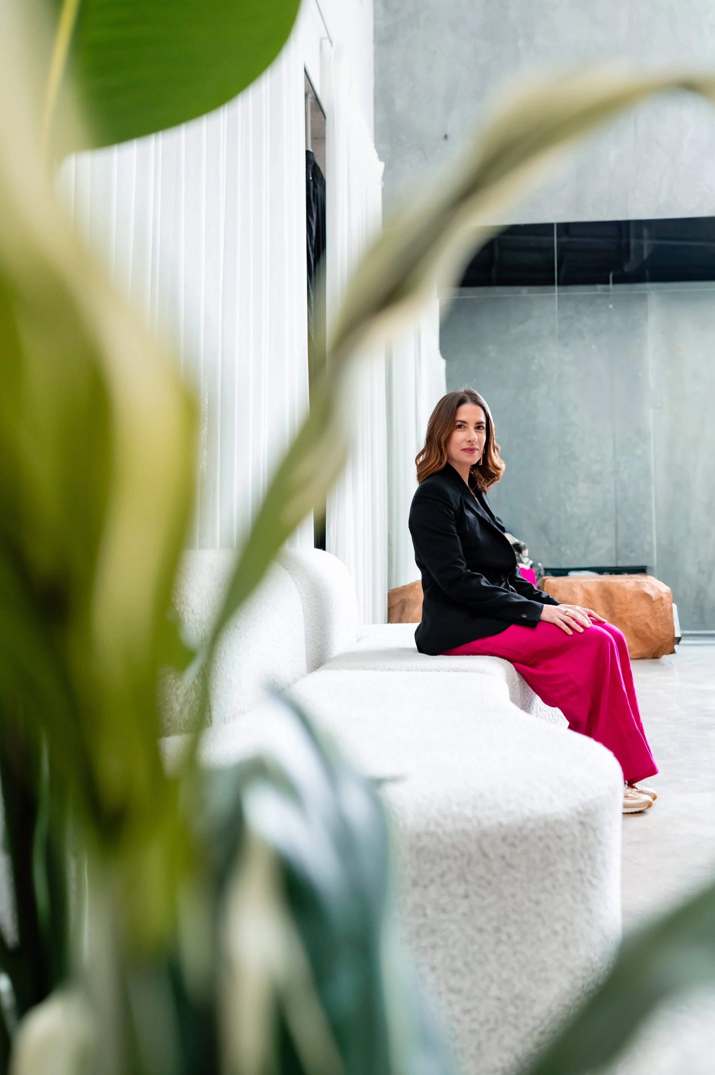 Woman in a black blazer and bright pink pants sitting on a white sofa in a modern living room