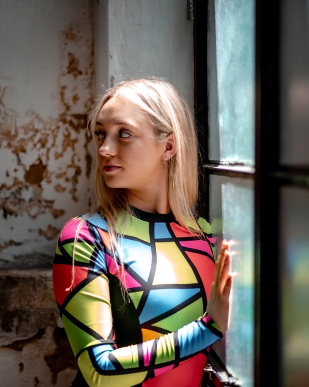 Blonde woman in a colorful geometric-patterned shirt standing beside a window in a weathered building