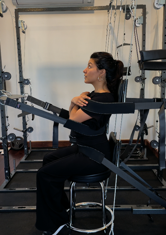 A woman sitting on a stool in a rehabilitation or physical therapy setting with an exoskeleton aid strapped to her body and arms, surrounded by exercise equipment.