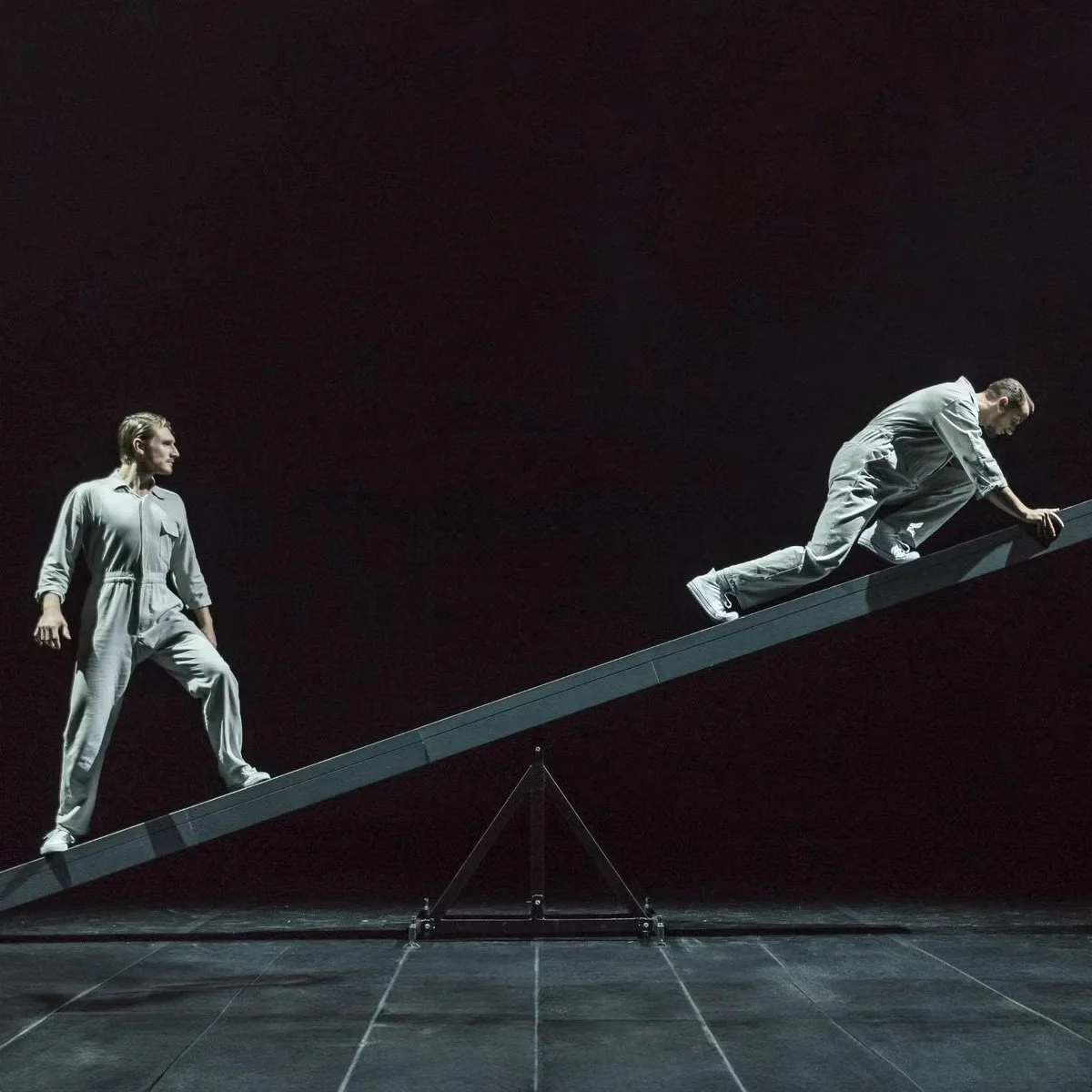 Two men dressed in gray outfits working together to walk up an inclined ramp in a dark space.