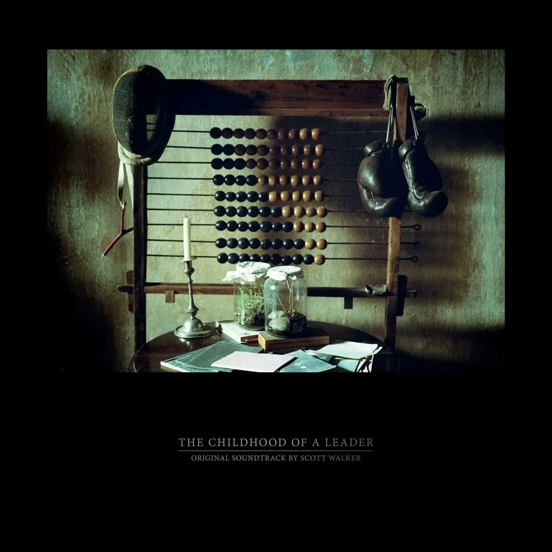 Album cover of 'The Childhood Of A Leader' featuring vintage boxing gloves hanging on a wooden frame with an abacus and a speaker, a table with a candlestick, jars, and papers against a textured wall.