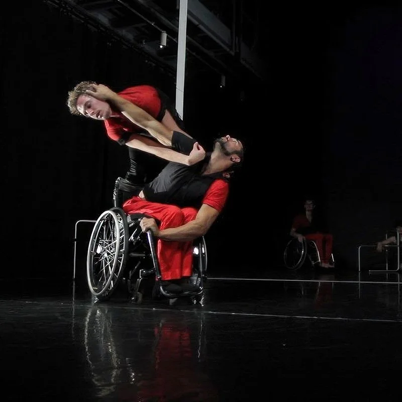 Two performers, one in a wheelchair, engaging in a dance or theatrical performance on a dark stage. The man in the wheelchair is holding the woman, who is leaning over him, as part of their act. Another person in a wheelchair is visible in the background.