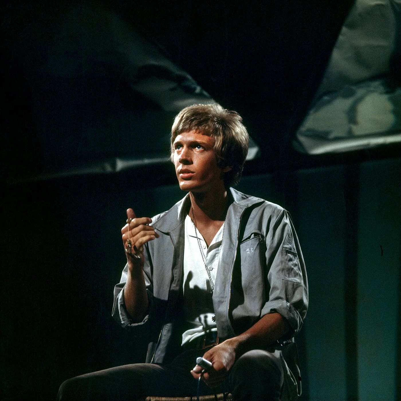 Scott Walker with a beaded necklace in his hand, sitting in a dimly lit TV studio with dark background, wearing a light gray jacket and a white shirt.