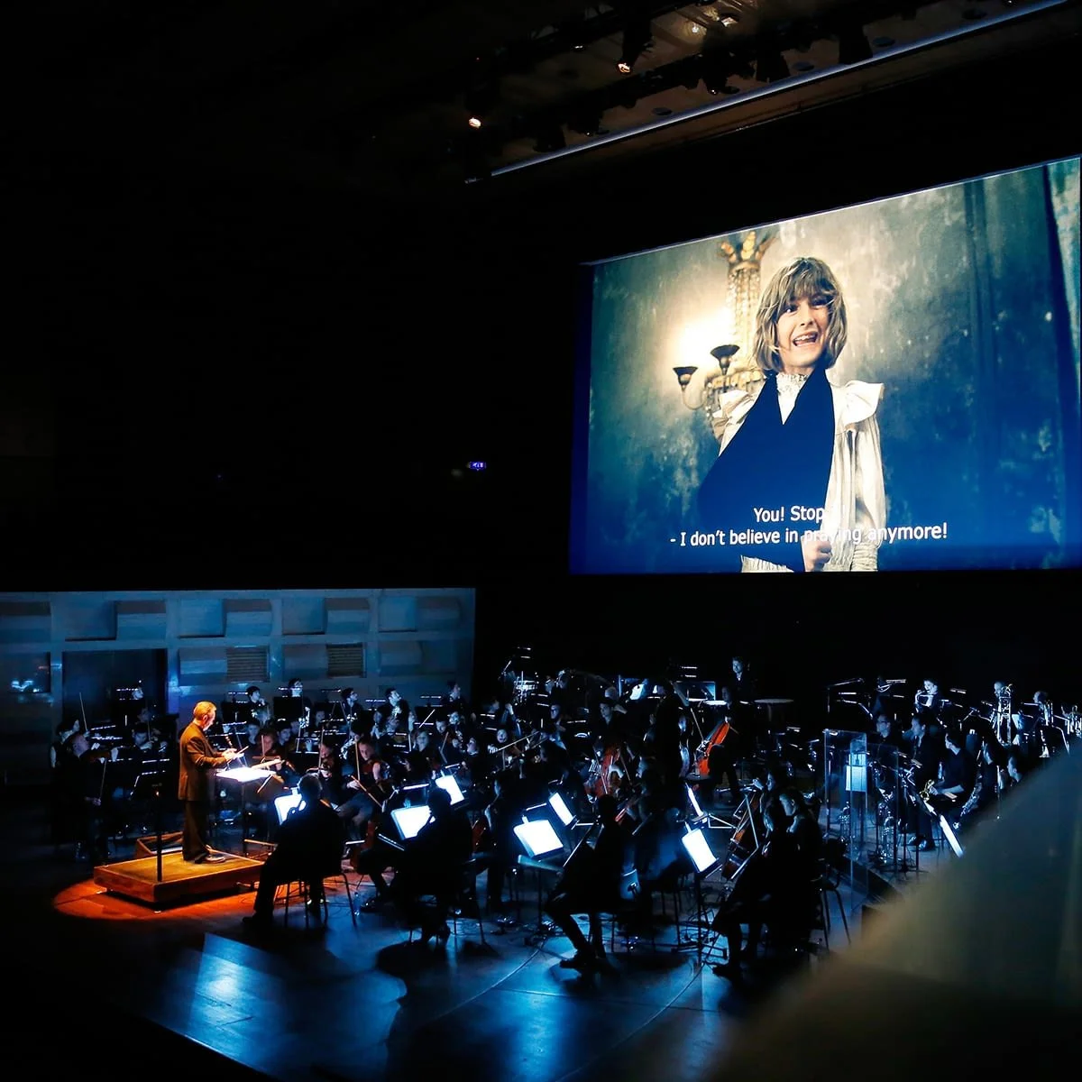 Orchestra performing on stage with a conductor, large screen above showing scene from the film 'The Childhood Of A Leader'.