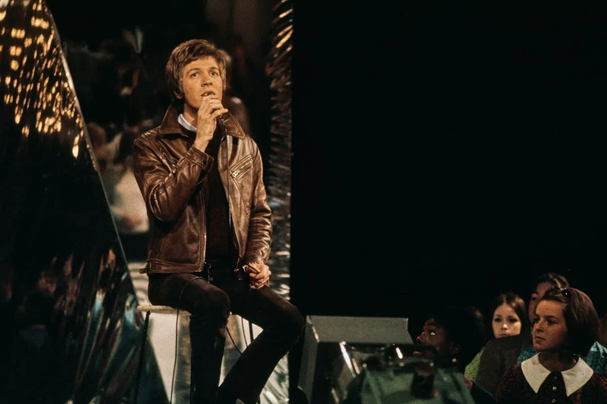 Scott Walker in a brown leather jacket singing into a microphone on a TV sound stage in front of an audience.