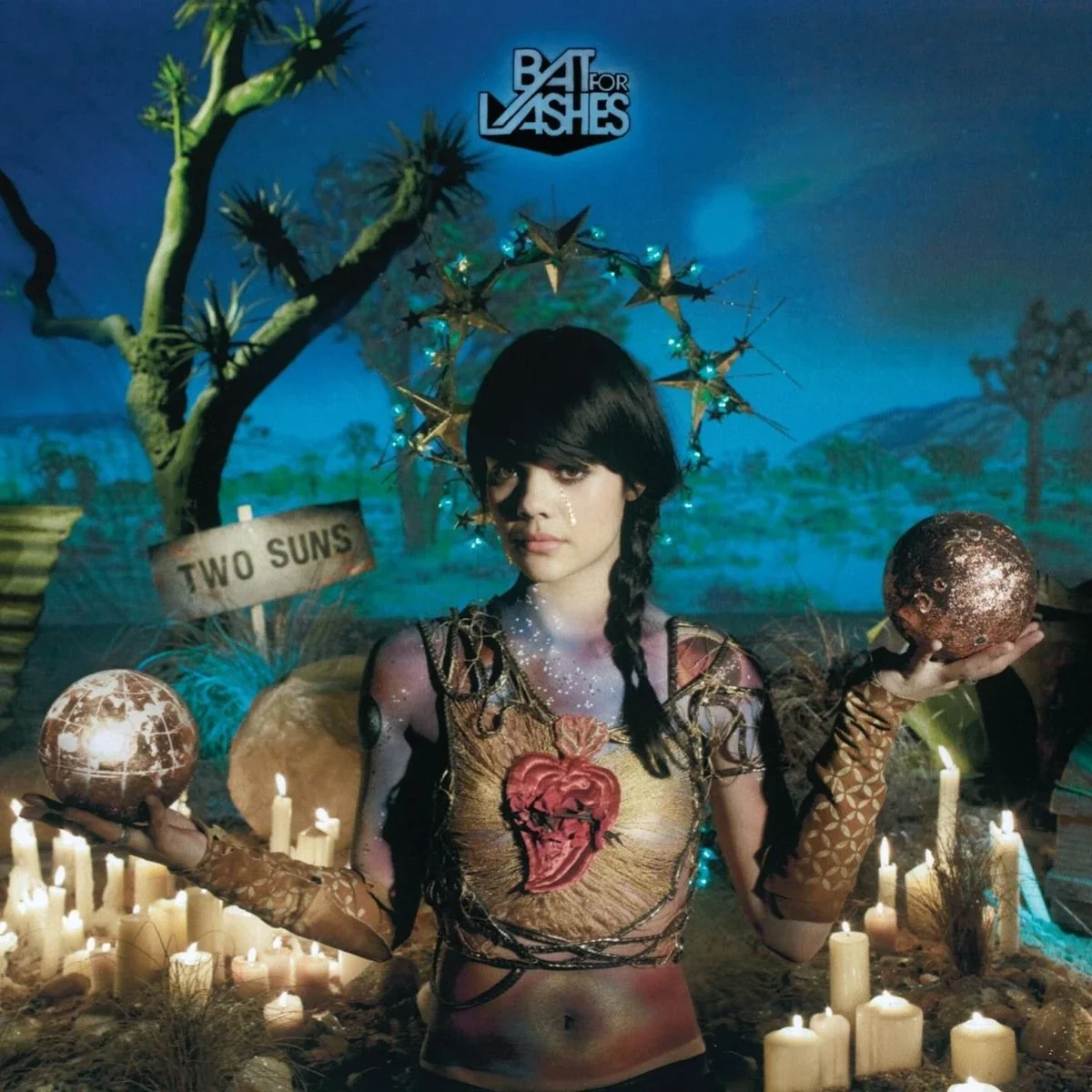 A woman with dark hair and a braid, dressed in a fantasy-themed costume with a heart design on her chest, is holding a glittery globe in each hand. She is surrounded by candles and planet-like objects. Behind her is a backdrop with a painted desert landscape and a sign that says "Two Suns." She has a star-shaped halo above her head and the words "BAT FOR LASHES" are displayed at the top.