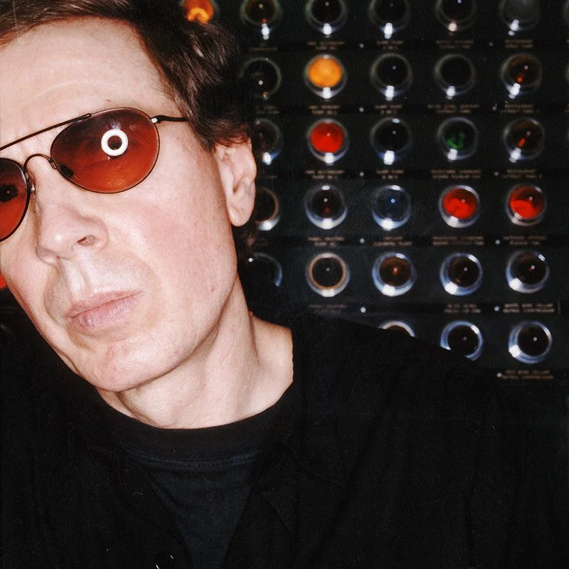 Portrait of Scott Walker wearing red-tinted sunglasses and a black shirt in front of a wall of colorful industrial console lights.