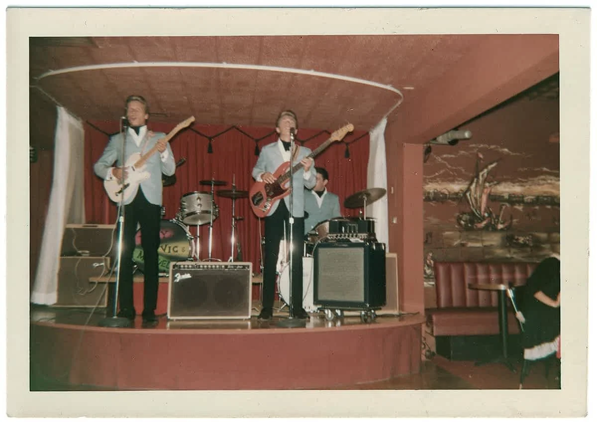 Scott on bass guitar, John playing electric guitar and Al Schneider playing drums on a small stage with red curtains (possibly Gazzarri's), The stage is decorated with Christmas lights and the band are dressed in light jackets and black pants.