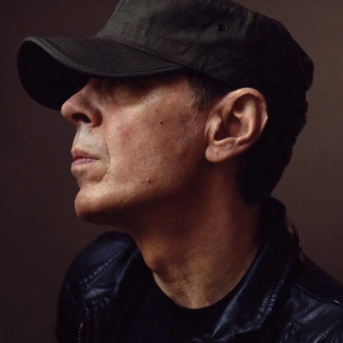 Close-up profile picture of a Scott Walker wearing a black military-style cap and a black leather jacket, with a serious expression.