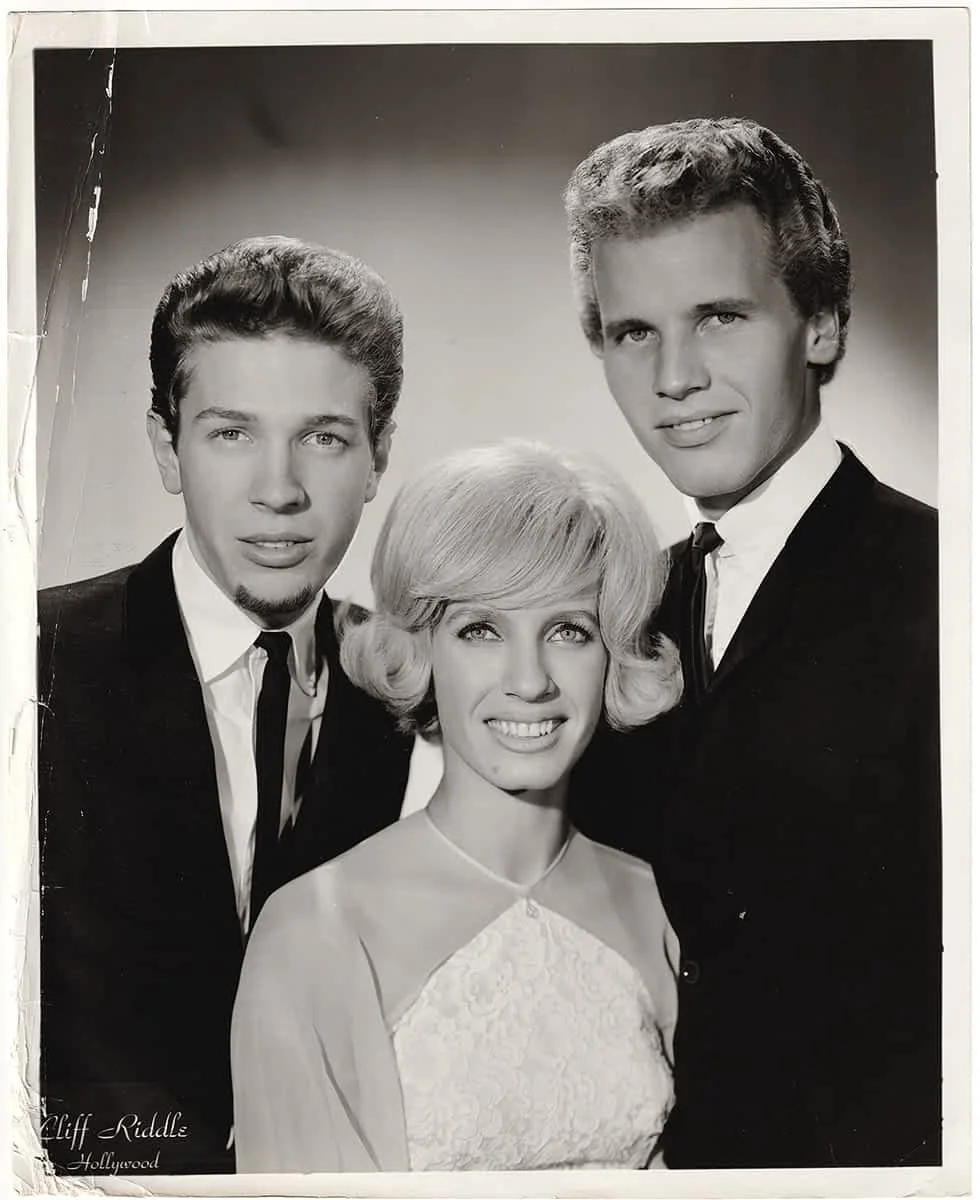 Posed black and white promotional portrait of Scott Walker, with Judy & John Maus dressed in formal attire, with a plain gradient background.