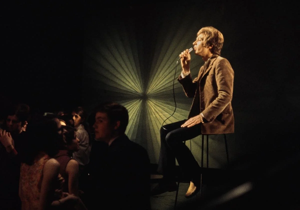 Scott Walker in a brown leather jacket singing into a microphone on a TV sound stage in front of an audience while seated on a high stool, with a dark backdrop featuring a large, radiant, starburst pattern.