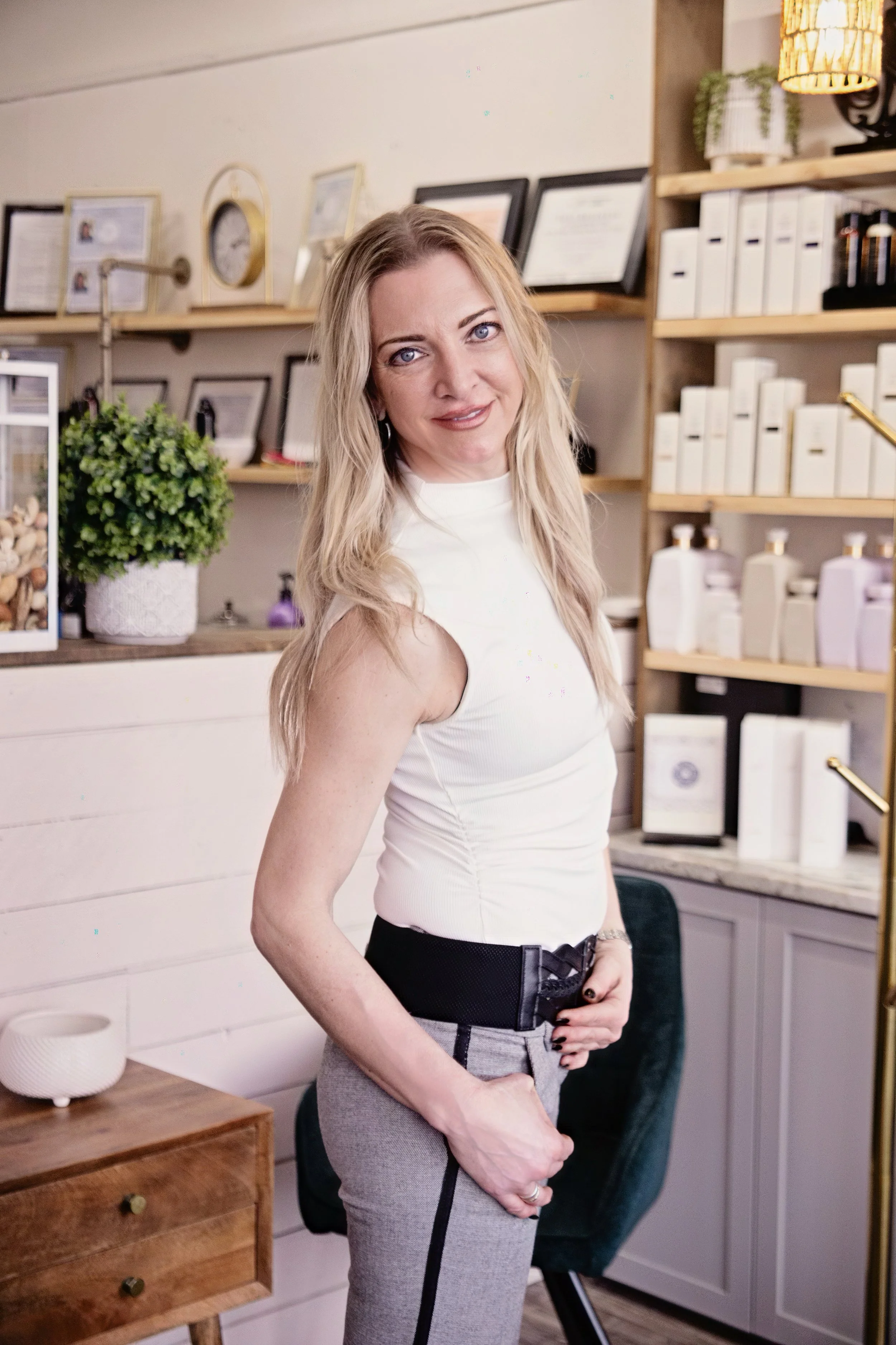 A woman with blonde hair, wearing a white sleeveless top and gray pants, stands in what appears to be a cozy, well-decorated room with shelves and framed pictures in the background.