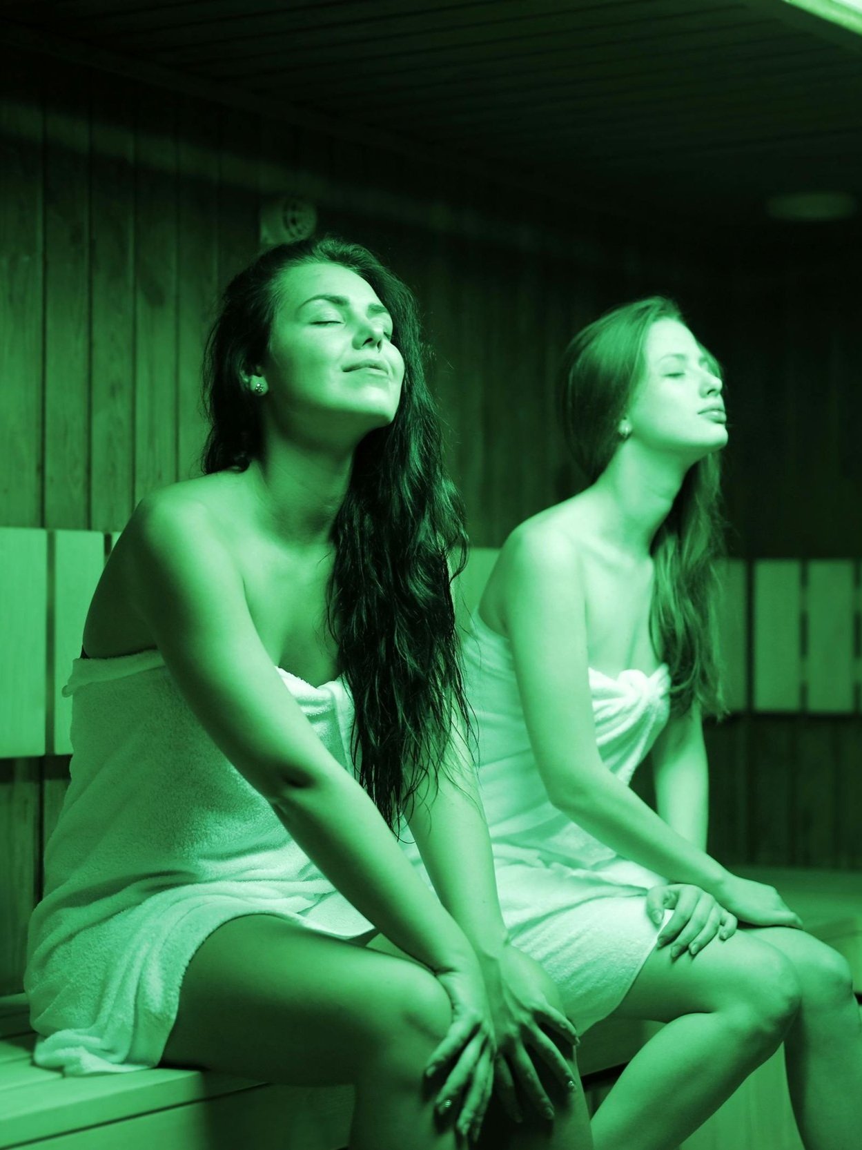 Two women sitting in a sauna, wrapped in towels, with eyes closed and relaxing.