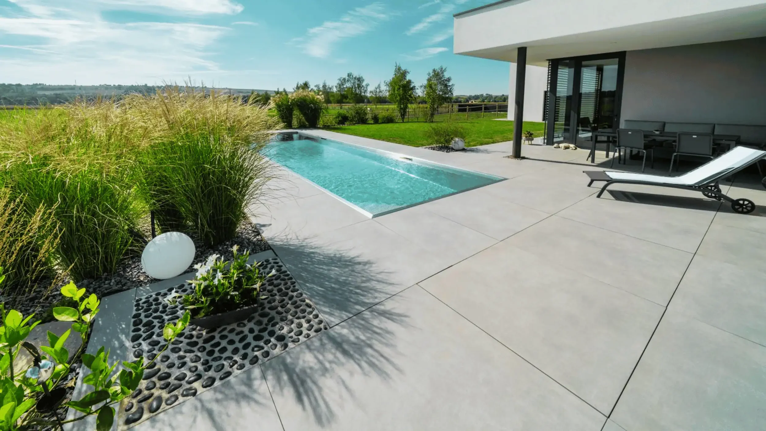 Moderne Terrasse mit Swimmingpool, Gartenmöbeln und Liegestuhl vor einem weißen Haus mit Balkon, unter blauem Himmel.