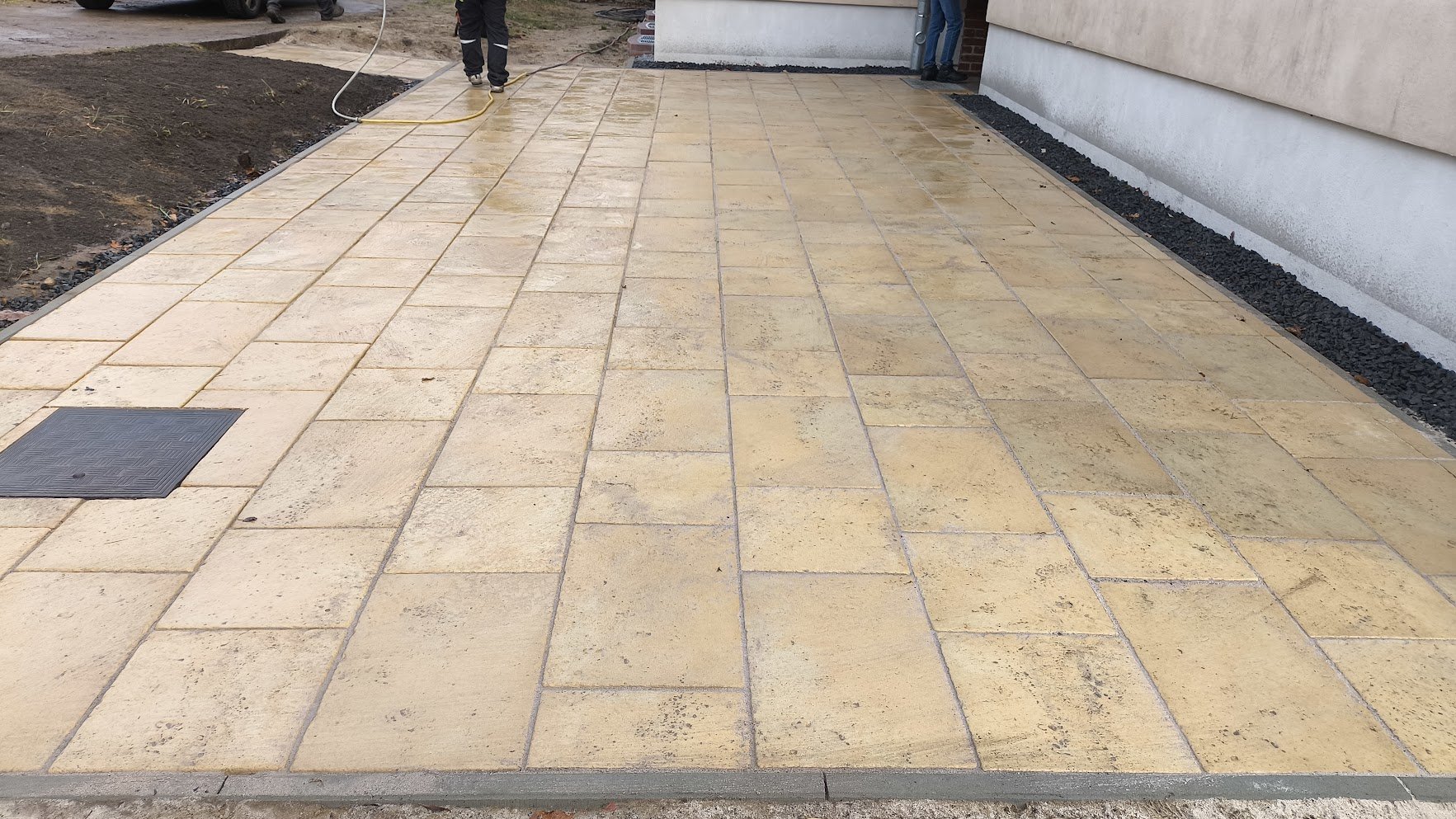 Neue Pflasterung auf Terrasse mit gelben Steinen, Seitenrand mit dunklen Kieselsteinen, im Hintergrund Bauarbeiter und Teil eines Hauses sichtbar.