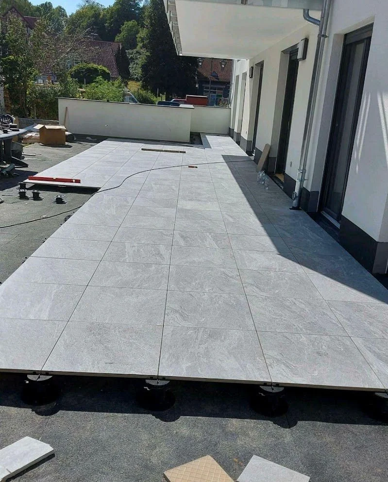 Balkon mit grauem Fliesenboden, im Hintergrund Gebäude und Bäume, Bauarbeiten mit Werkzeugen und Material ohne Geländer.