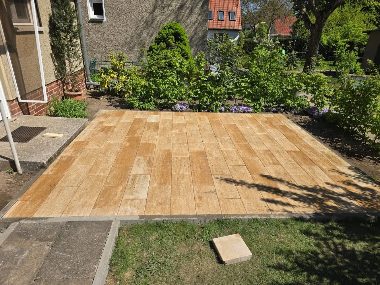 Garten mit Holz-Terrasse, umgeben von grünen Pflanzen und Bäumen bei Sonnenschein.