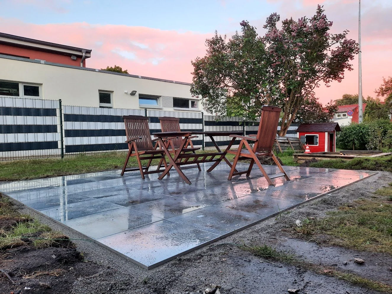 Essbereich im Garten mit vier aus Holz gefertigten Stühlen und einem Tisch auf nassen Fliesen, im Hintergrund ein Baum, eine Hundehütte und ein Zaun, bei Sonnenuntergang