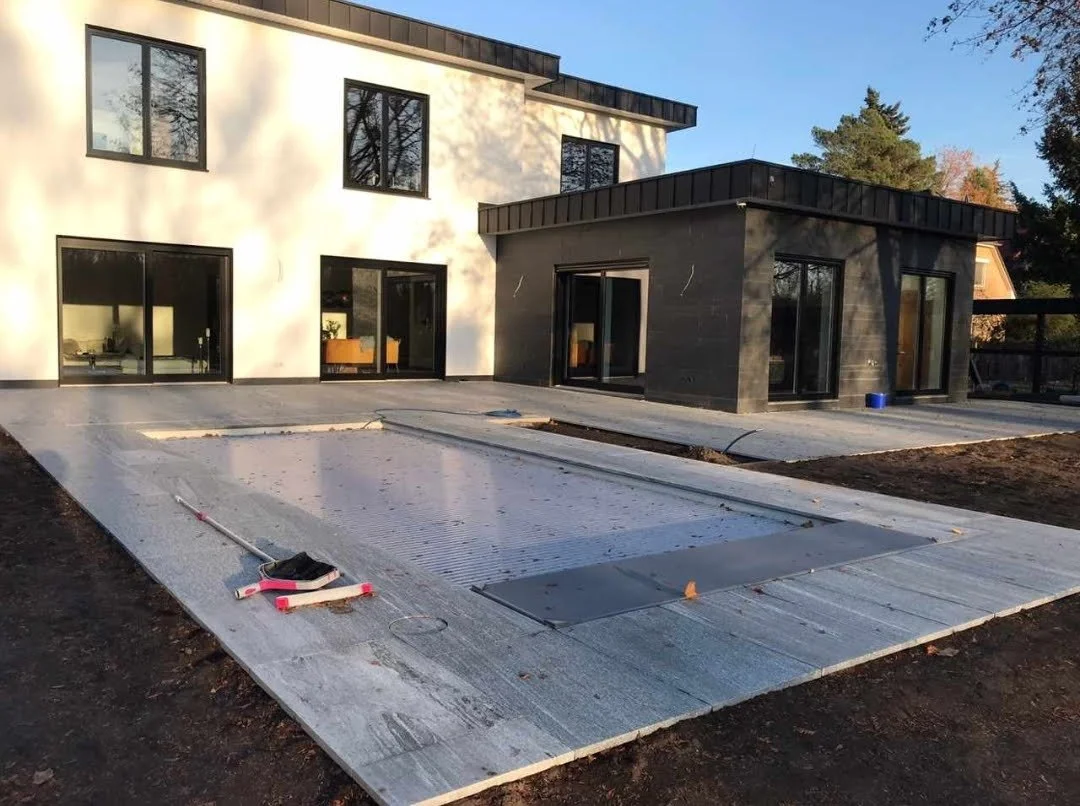 Ein modern gebautes Zweifamilienhaus mit großen Fenstern, im Vordergrund befindet sich eine noch im Bau befindliche Swimmingpool-Abdeckung.