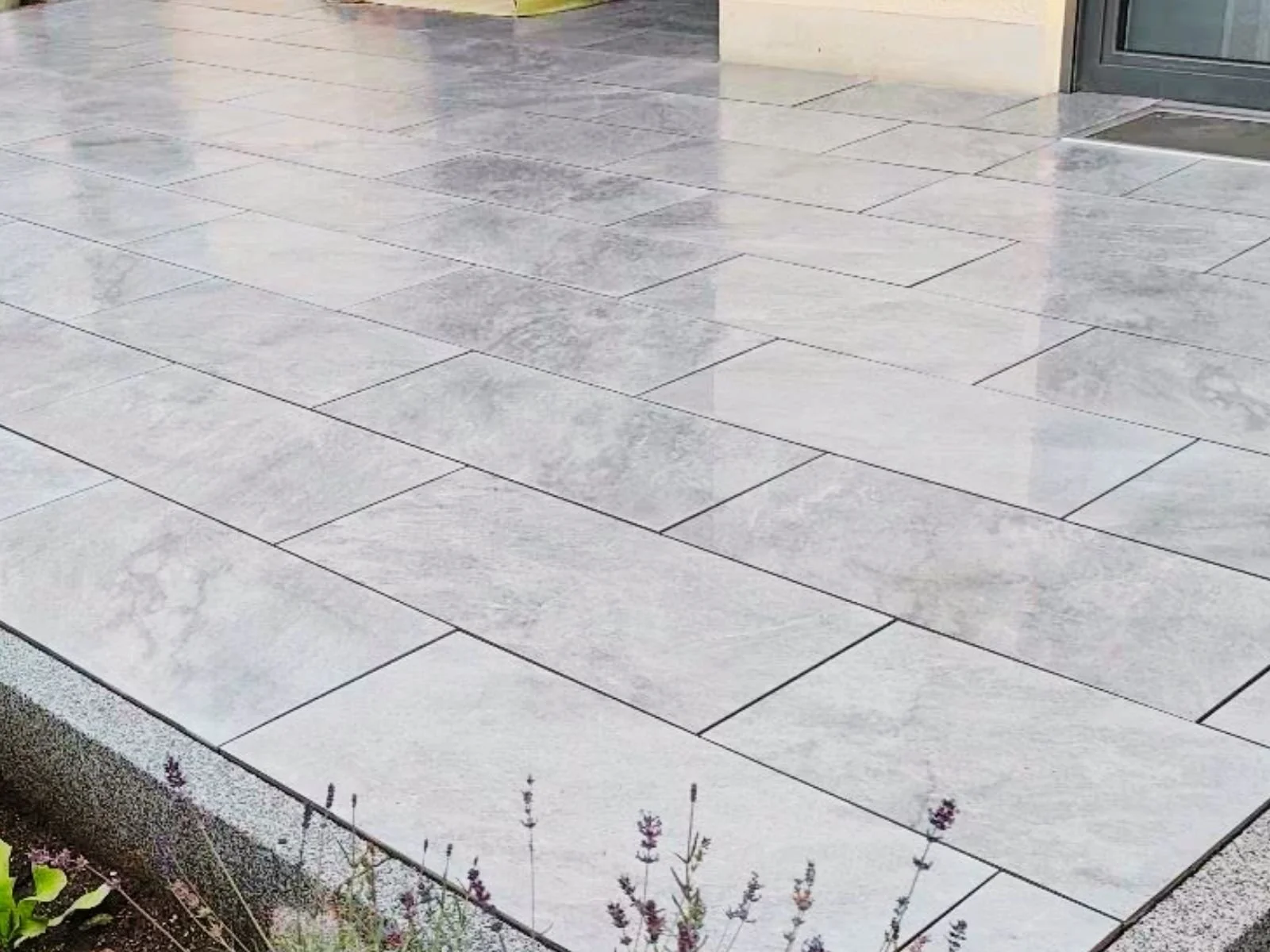 Blick auf einen gepflasterten Gehweg mit hellgrauen Steinen vor einem Geschäft mit Glastür und -fenster.