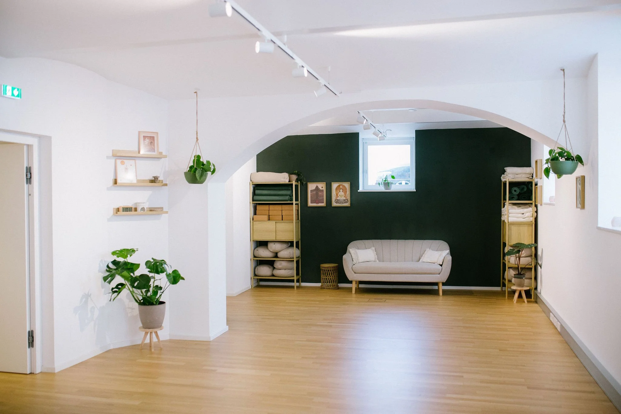 Ein hell erleuchteter Raum mit Holzfußboden, weißer Wände und einer dunklen Akzentwand. Es stehen Pflanzen, Regale mit Handtüchern und Dekorationen, sowie eine Couch.