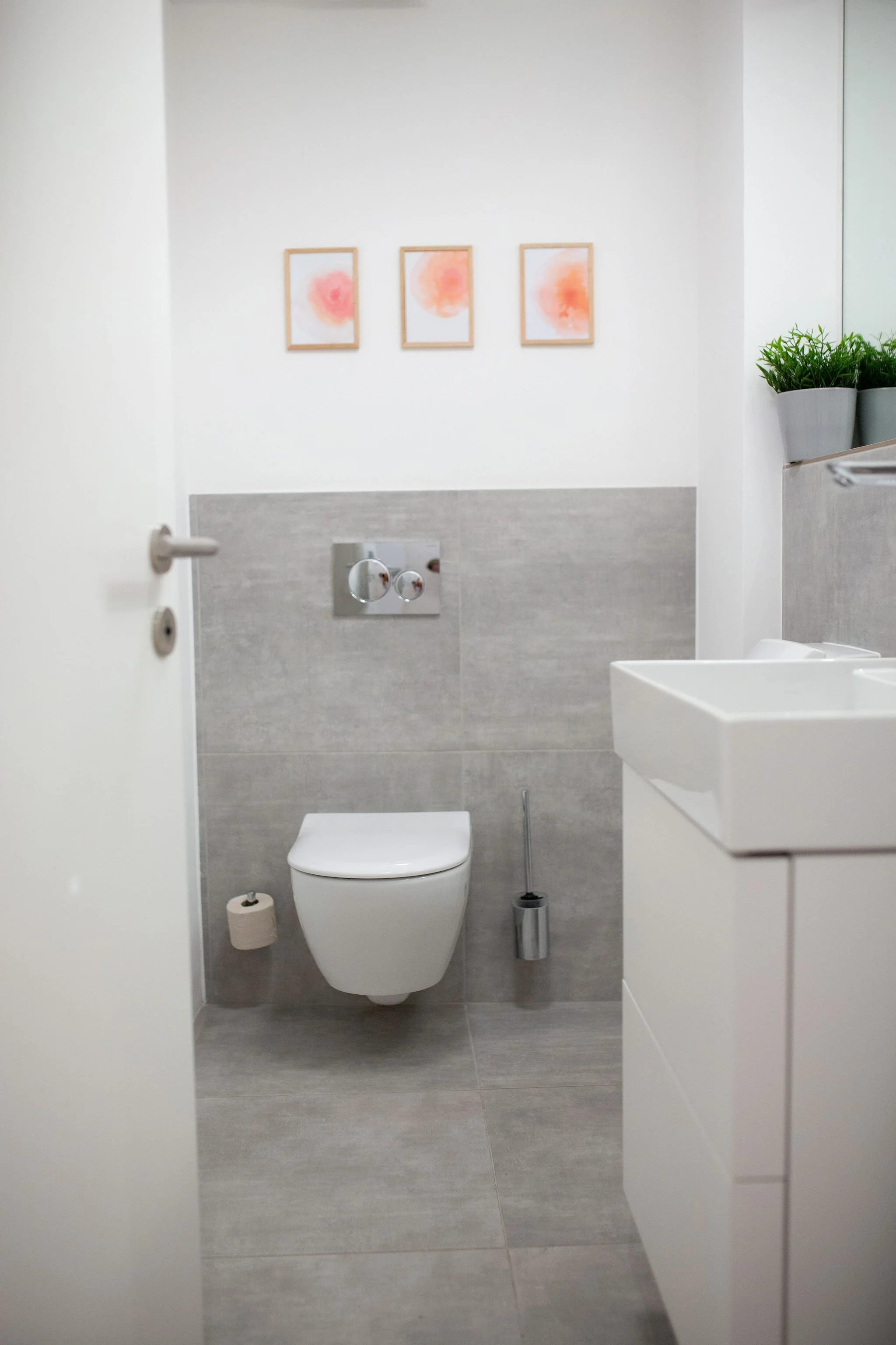 Kleines weißes Badezimmer mit einer Wand-Toilette, einem weißen Waschbecken, zwei Bilder mit roten Wasserfarben an der Wand, einem dutschenden Toilettenbürste und einer kleinen Pflanze auf einem Fensterbrett.