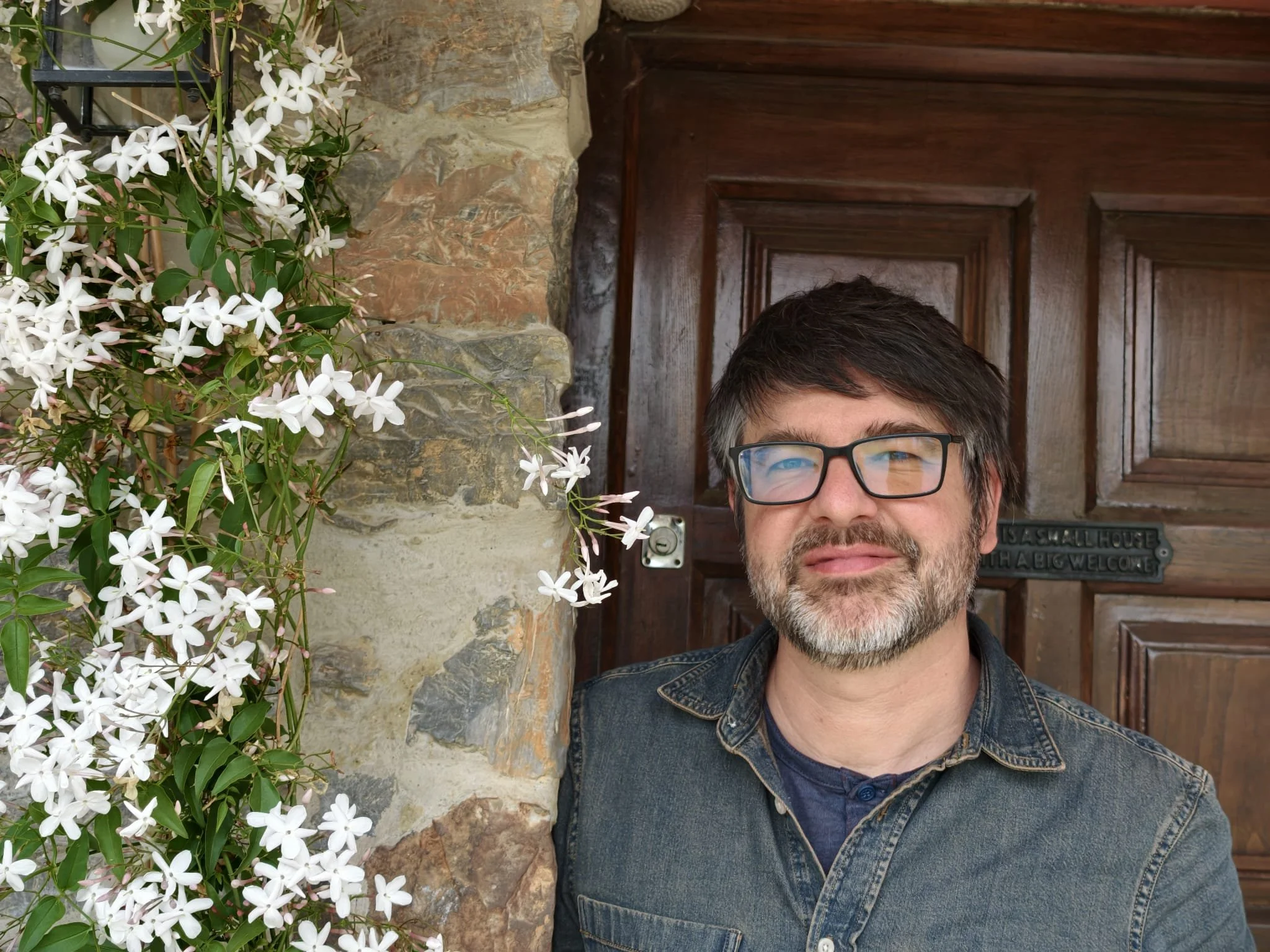 A man with glasses and a beard smiling in front of a wooden door and stone wall, with white flowers and green leaves in the foreground.